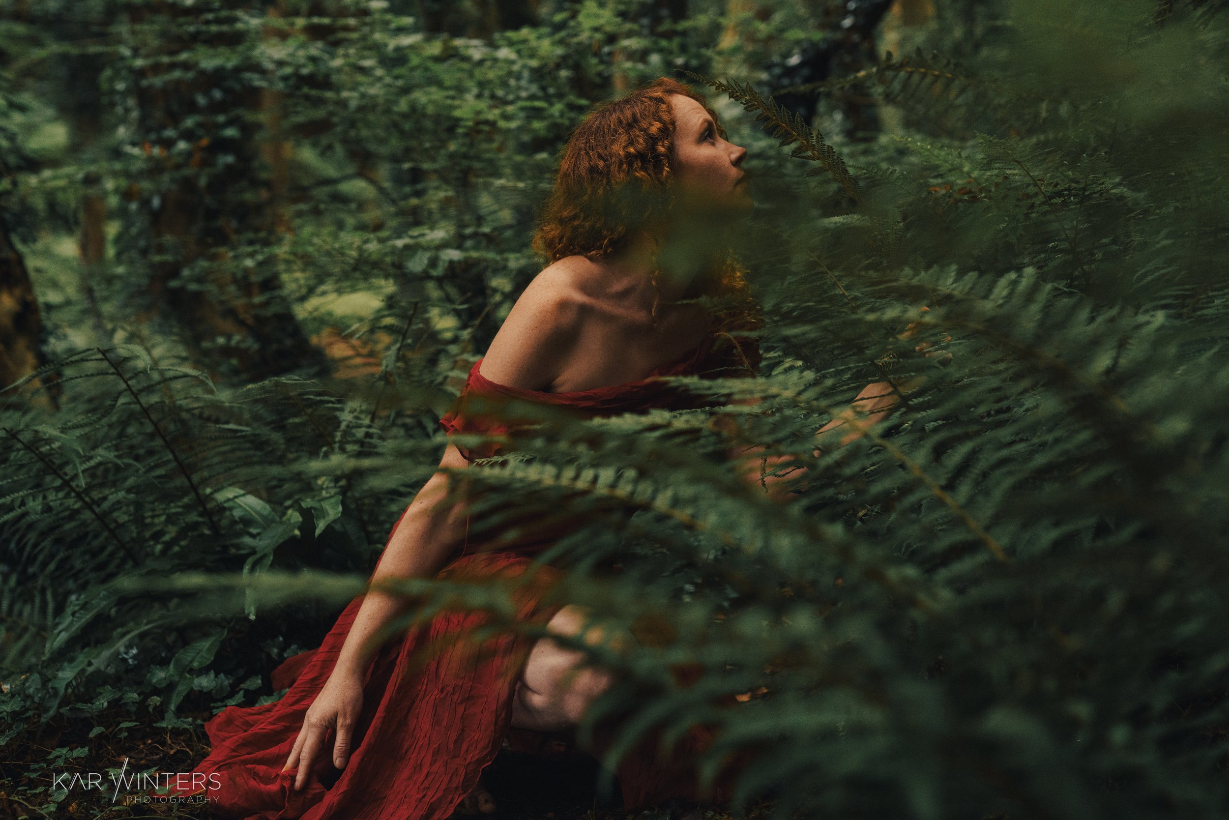 A woman with curly red hair in a red dress crouches in a lush, green forest with dense foliage and ferns, looking upward.