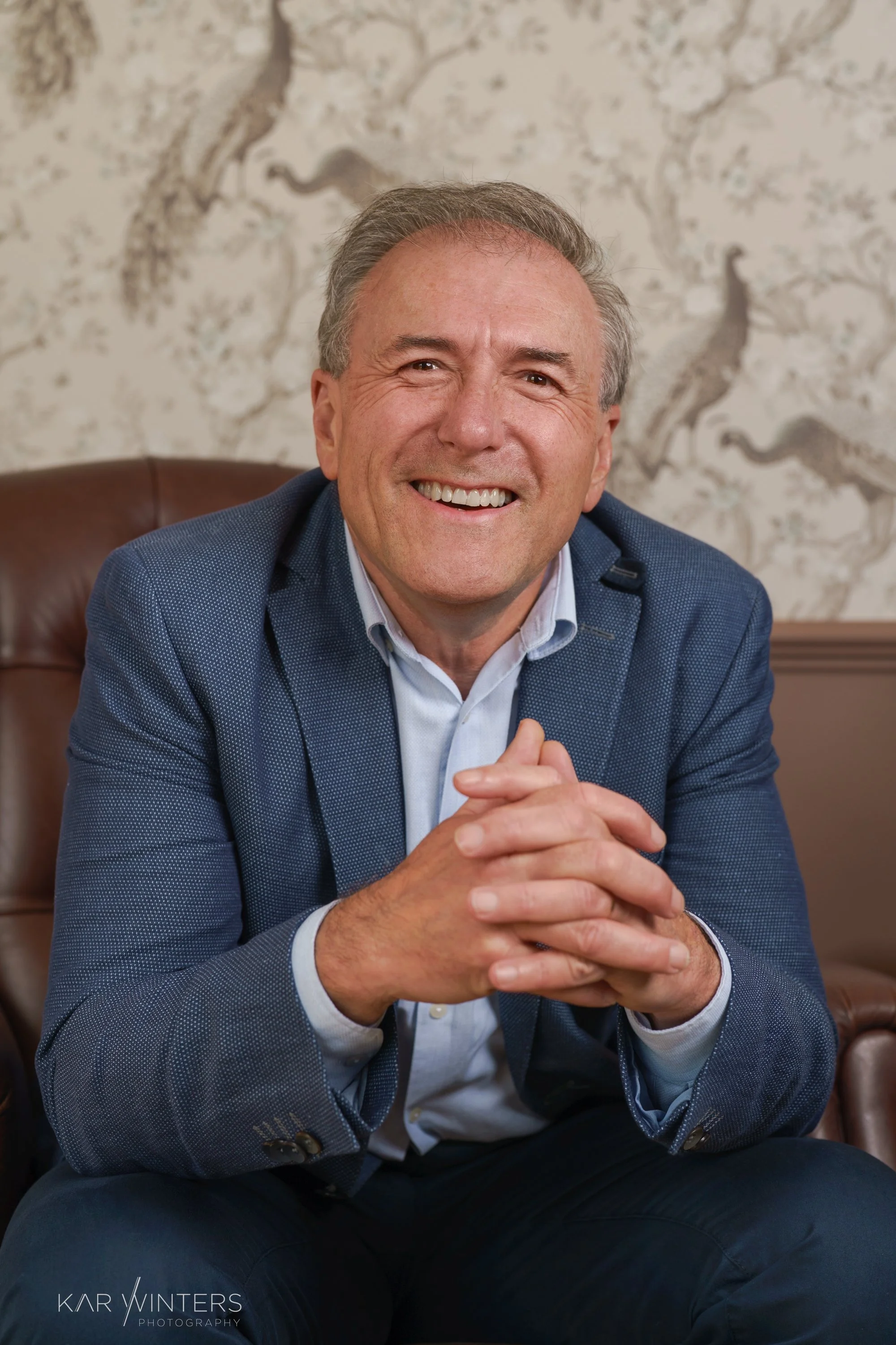 A smiling man sitting on a leather chair, wearing a blue blazer and a white shirt, with patterned wallpaper featuring birds in the background.
