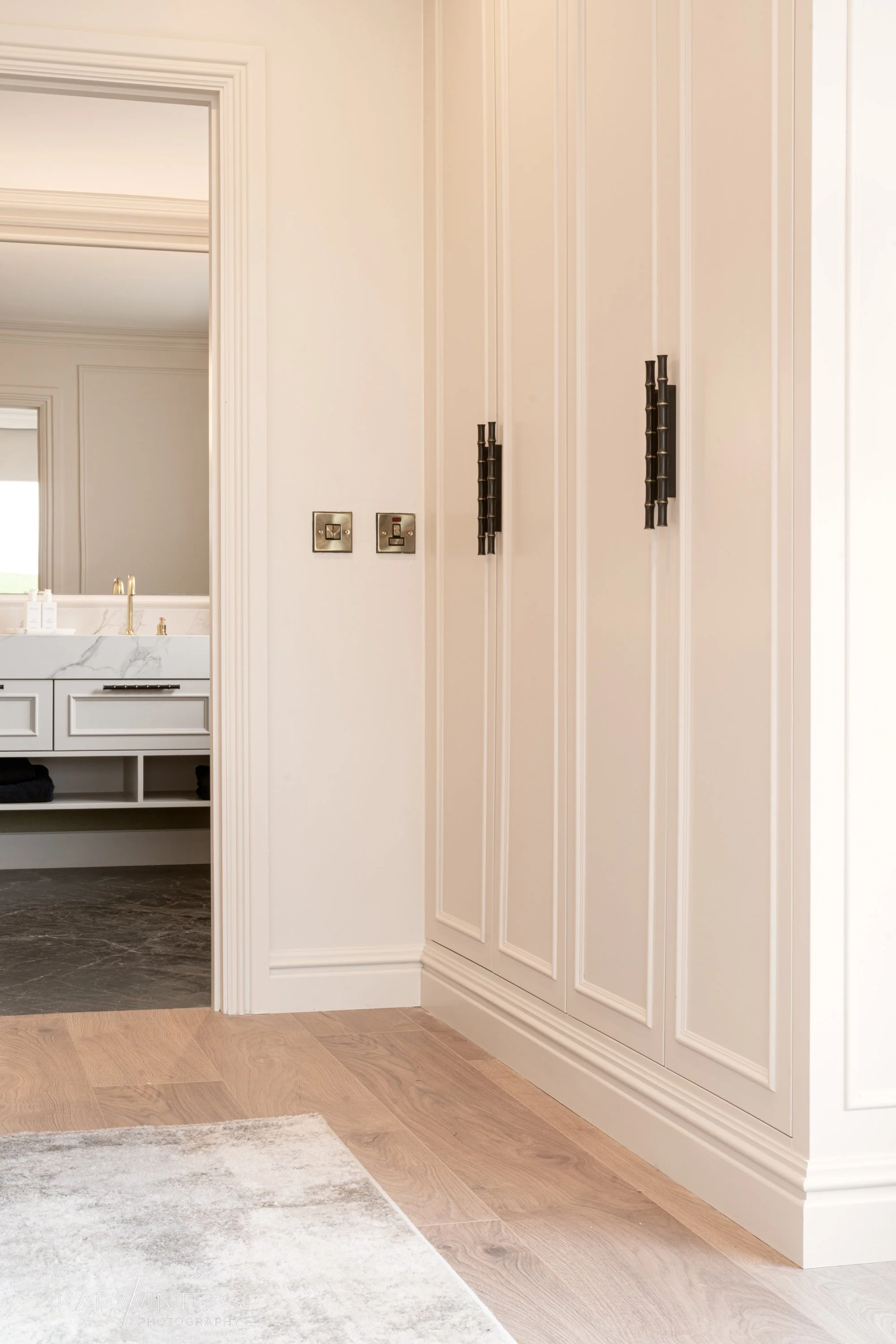 Interior view of a closet with black handles on white paneled doors, adjacent to a bathroom with a marble countertop sink and a large mirror, on light wood flooring and a plush beige rug.
