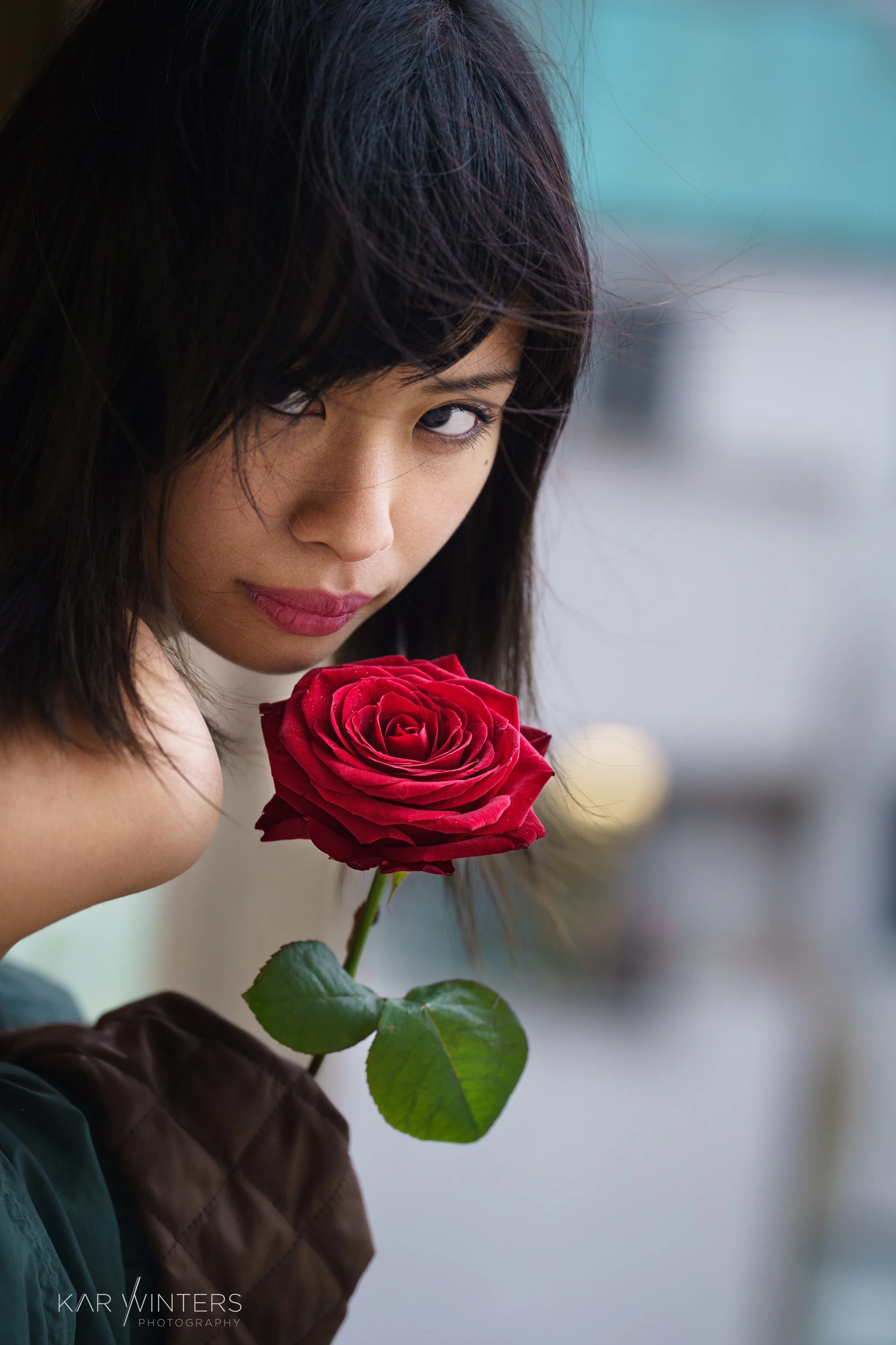 A woman with dark hair and makeup holding a red rose close to her face, looking directly at the camera.