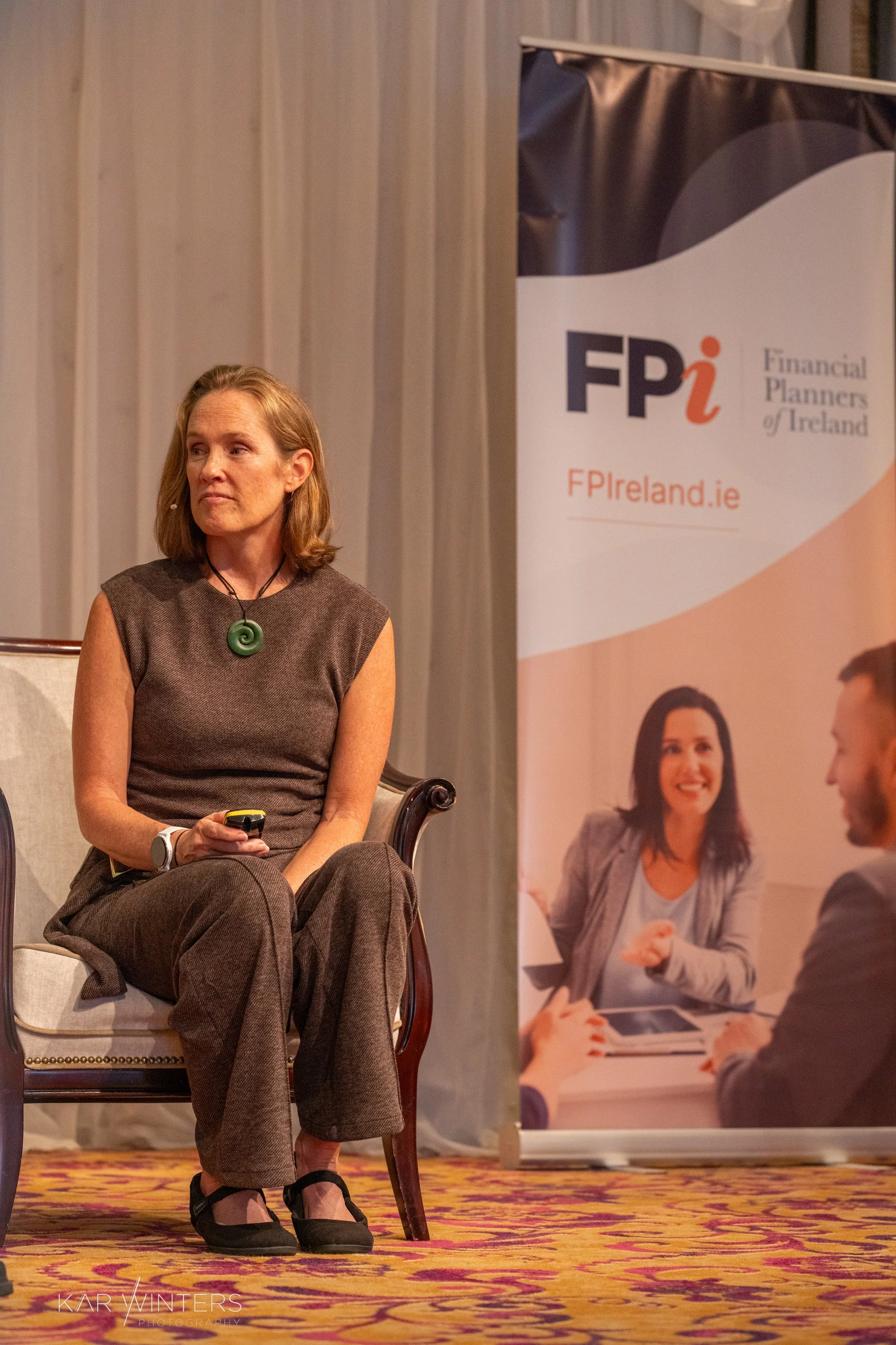 A woman sitting on a beige chair holding a remote during a professional event, with a FP Ireland banner beside her featuring a group of people in a business meeting.