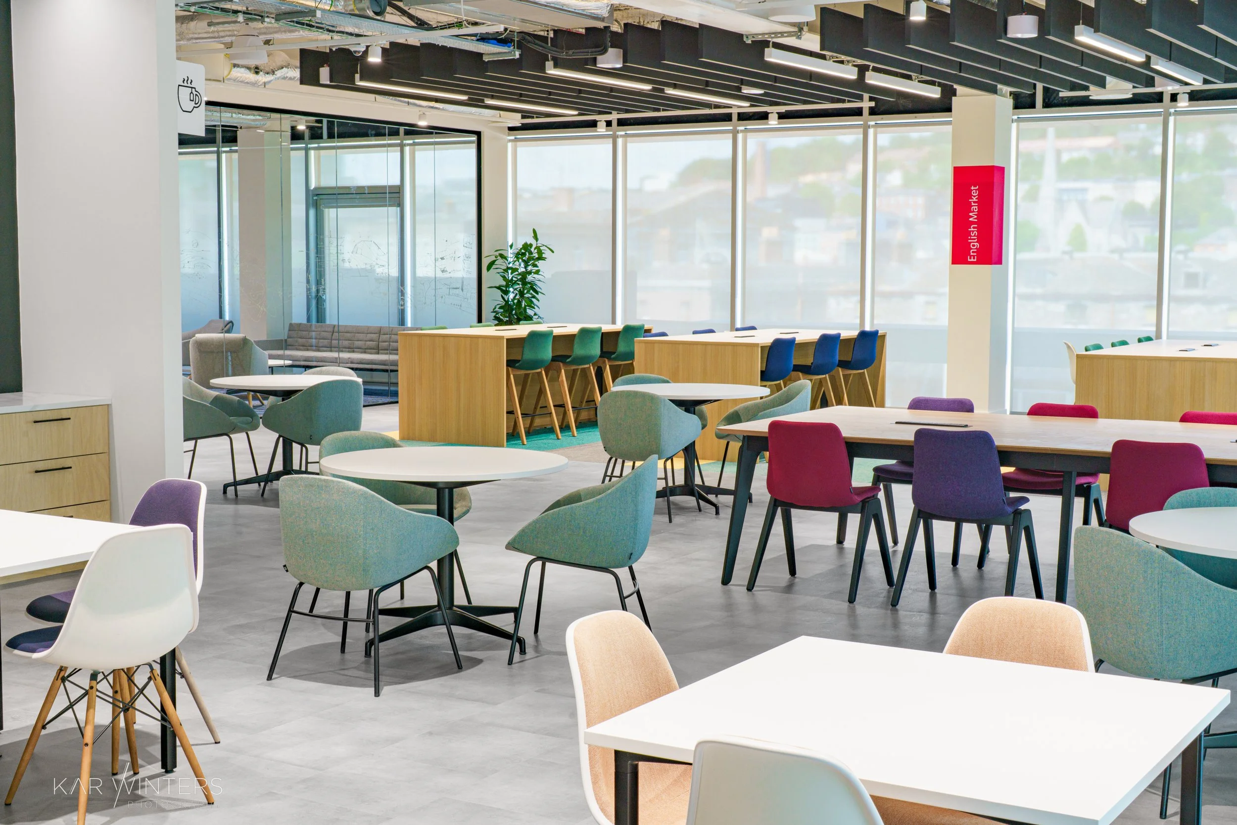 Modern, brightly lit cafe or communal workspace with colorful chairs, white tables, large windows, and a seating area with a sofa and a potted plant.