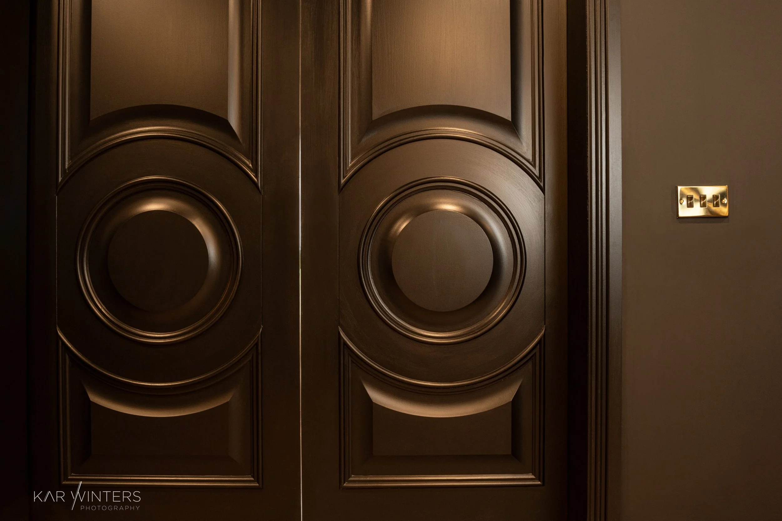 Close-up of dark wooden double doors with decorative circular carvings and a light switch on the tan wall to the right.