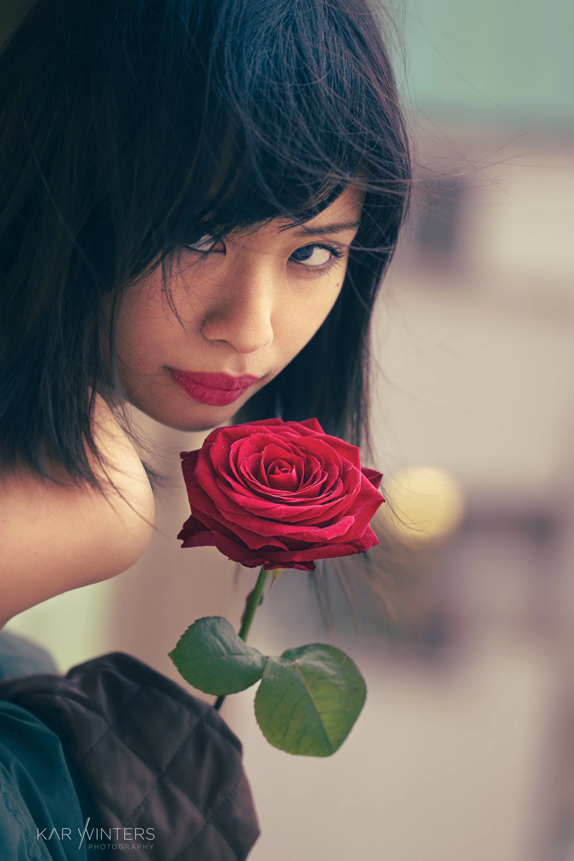 A woman with dark hair and red lipstick holding a red rose near her face and gazing at the camera.