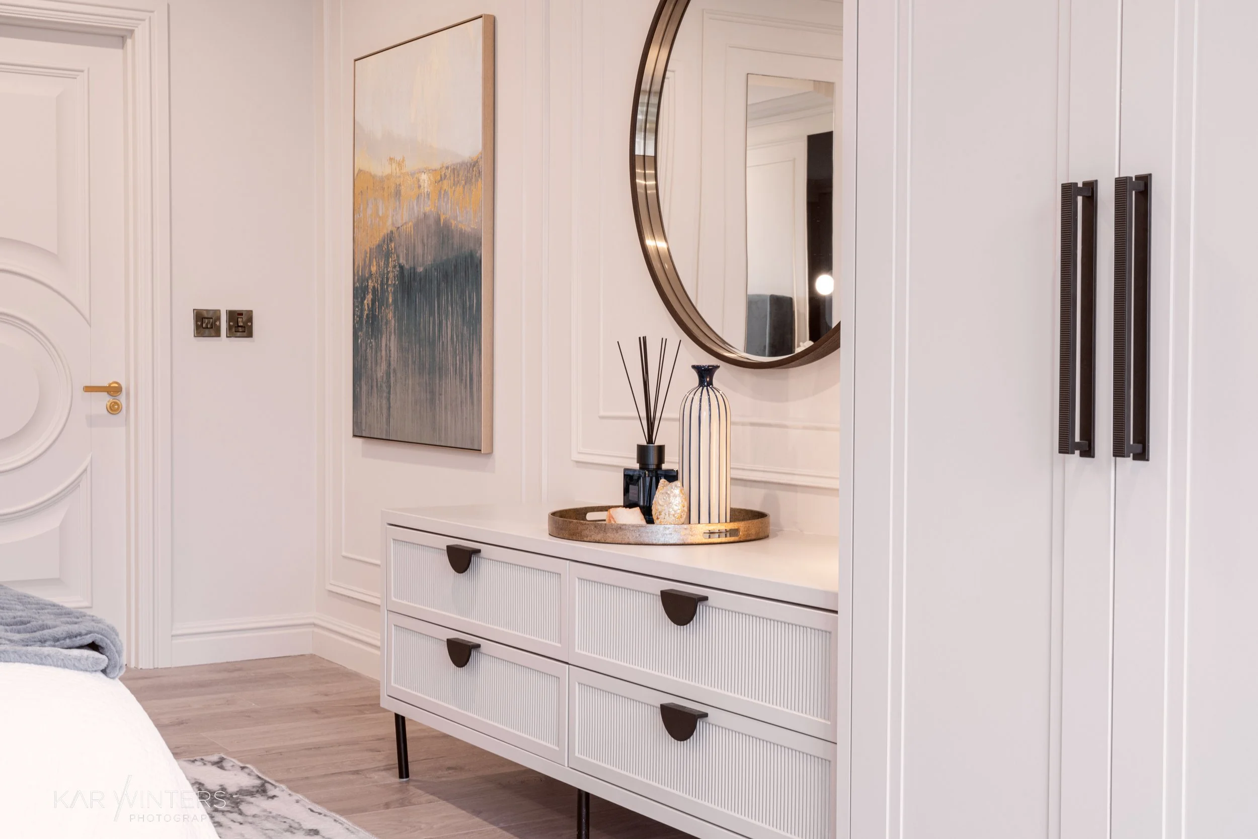 A modern bedroom dresser with decorative items, a round mirror, and wall art in a bright, minimalist interior.
