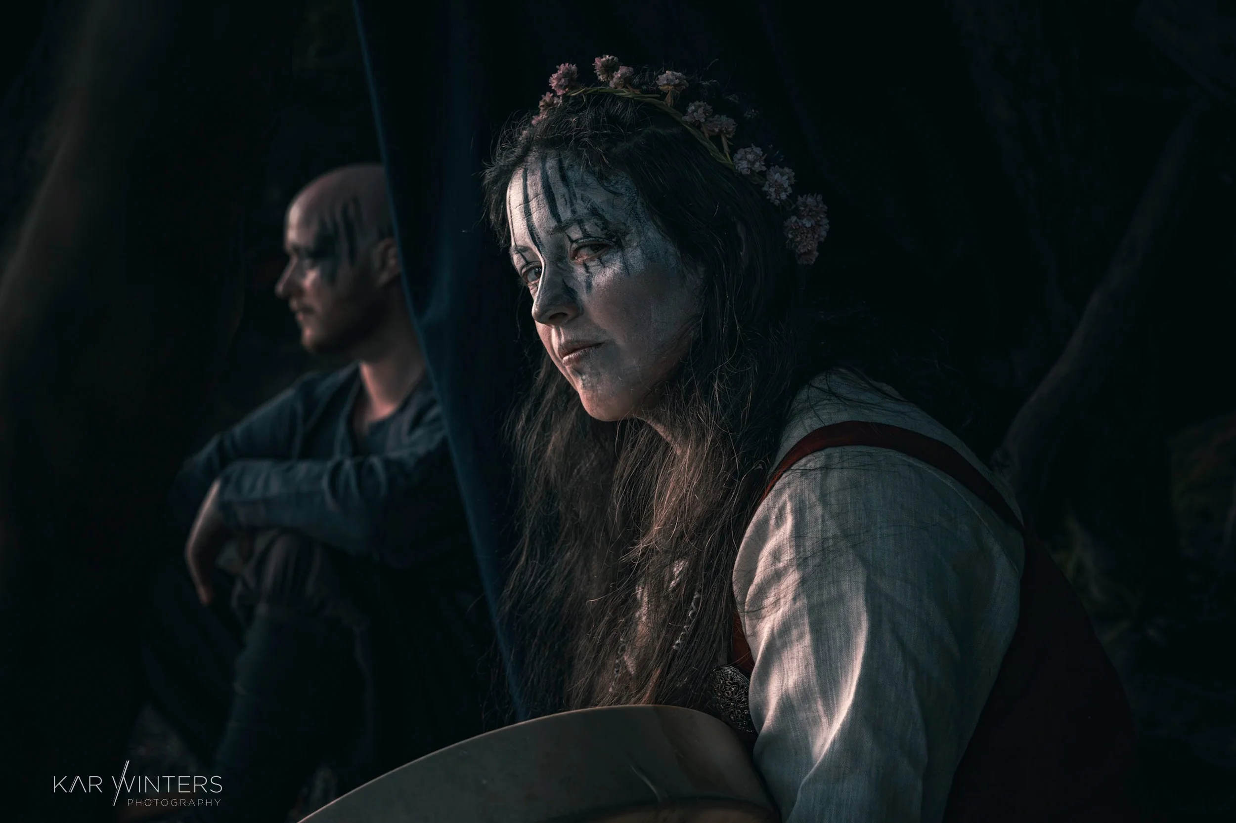 A woman with face paint, long hair, and a floral crown sitting in a dark space, with a man with glasses and short hair sitting behind her, both appearing somber.