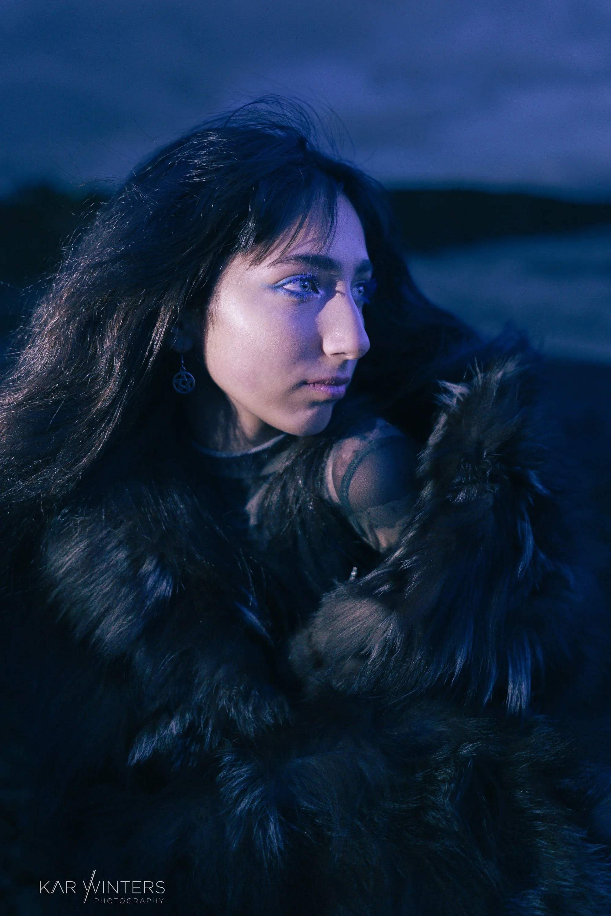 A woman with long dark hair and blue contact lenses wearing a fur coat and black top, looking to the side, outdoors at night with a blurred dark landscape in the background.