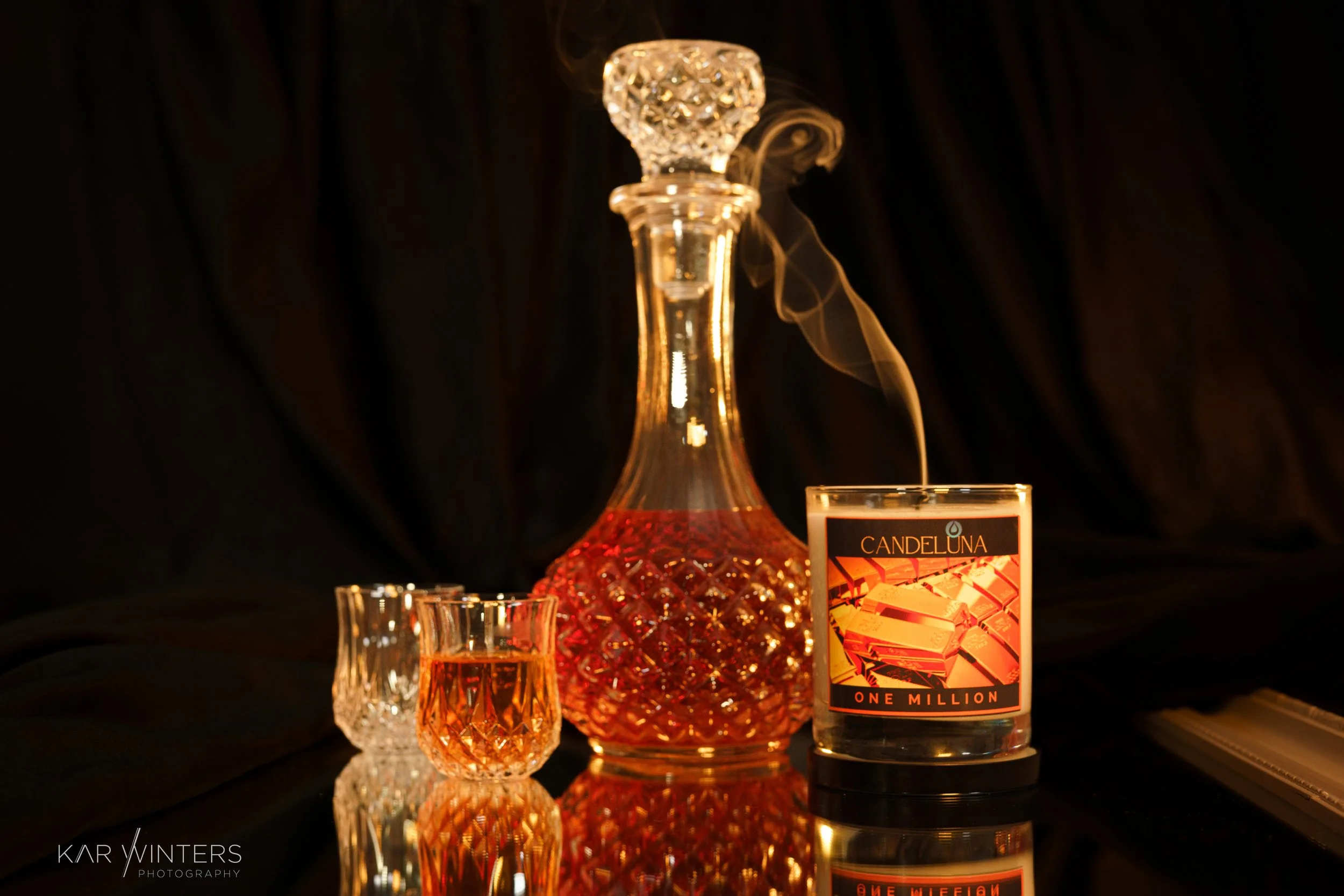 A lit candle, a glass decanter filled with amber liquid, and two small matching glasses of the same liquid are on a reflective black surface. The candle has a label that reads "Candeluna One Million" with an image of gold bars. The background is dark