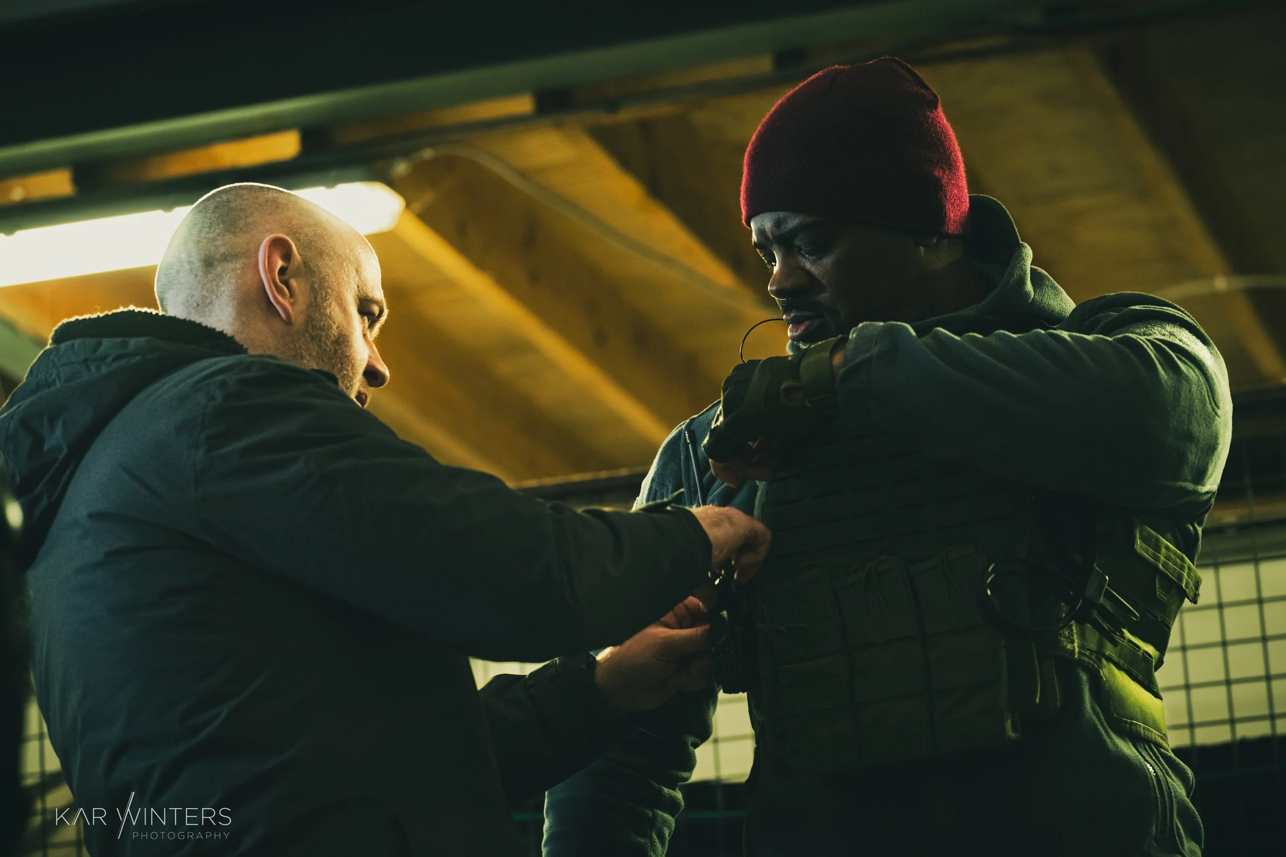 A person in tactical gear adjusting their uniform or gear while another person assists, in an indoor setting with wooden ceiling panels.