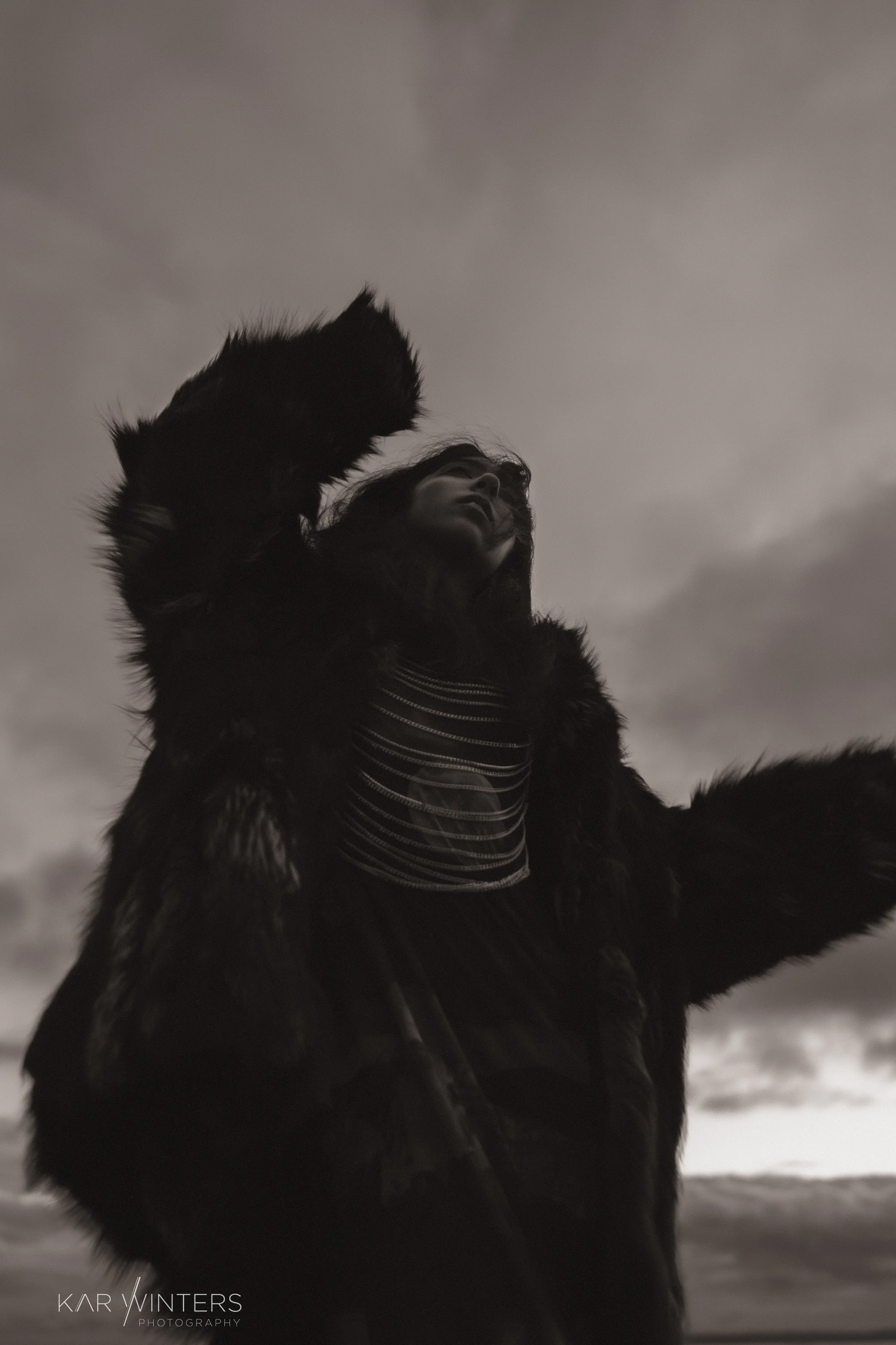 A black and white photo of a person wearing a fur coat and striped shirt, looking upwards with one arm raised, against a cloudy sky.