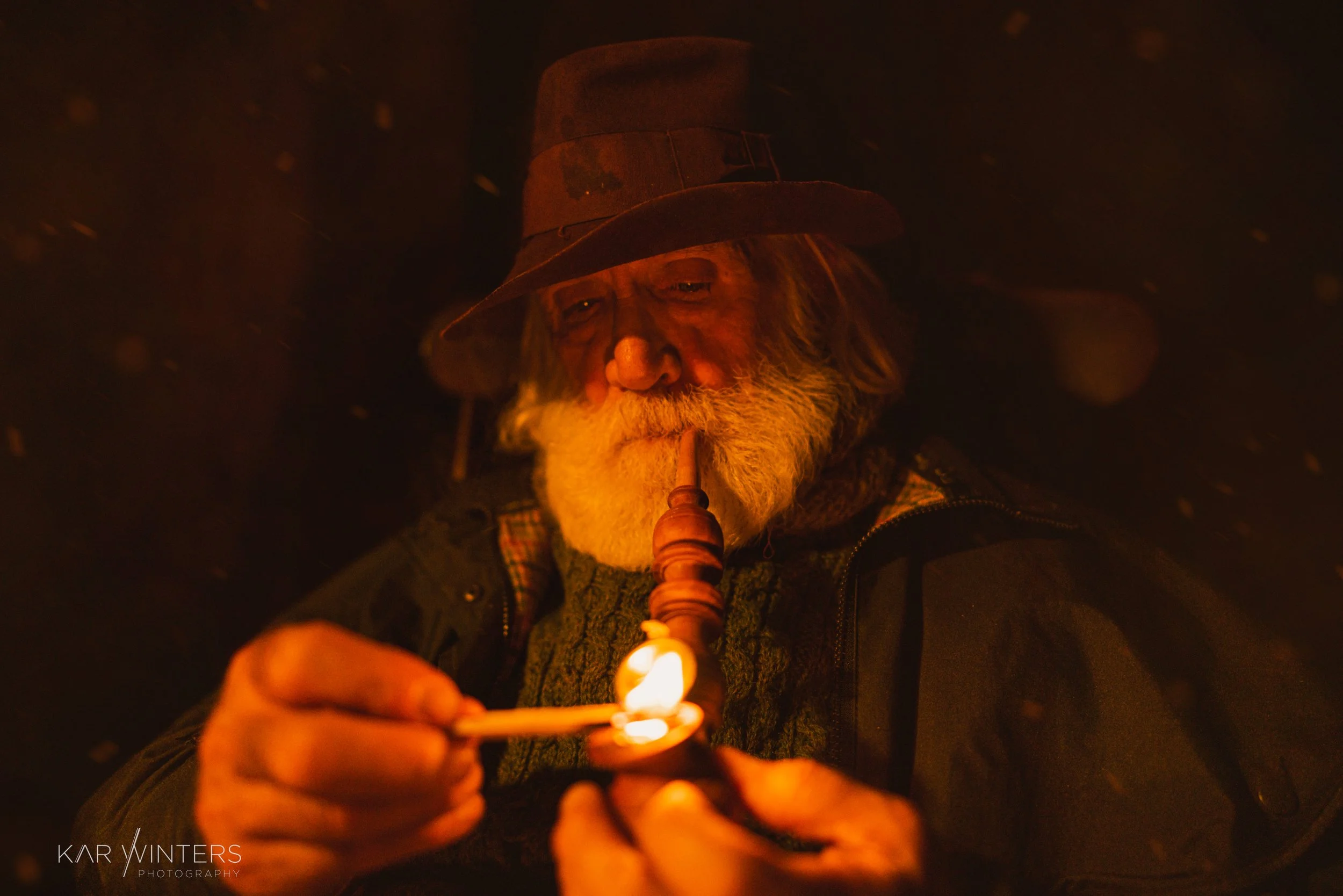 An elderly man with a white beard, wearing a hat, is lighting a pipe with fire from a match in a dark setting with glowing embers or particles around him.