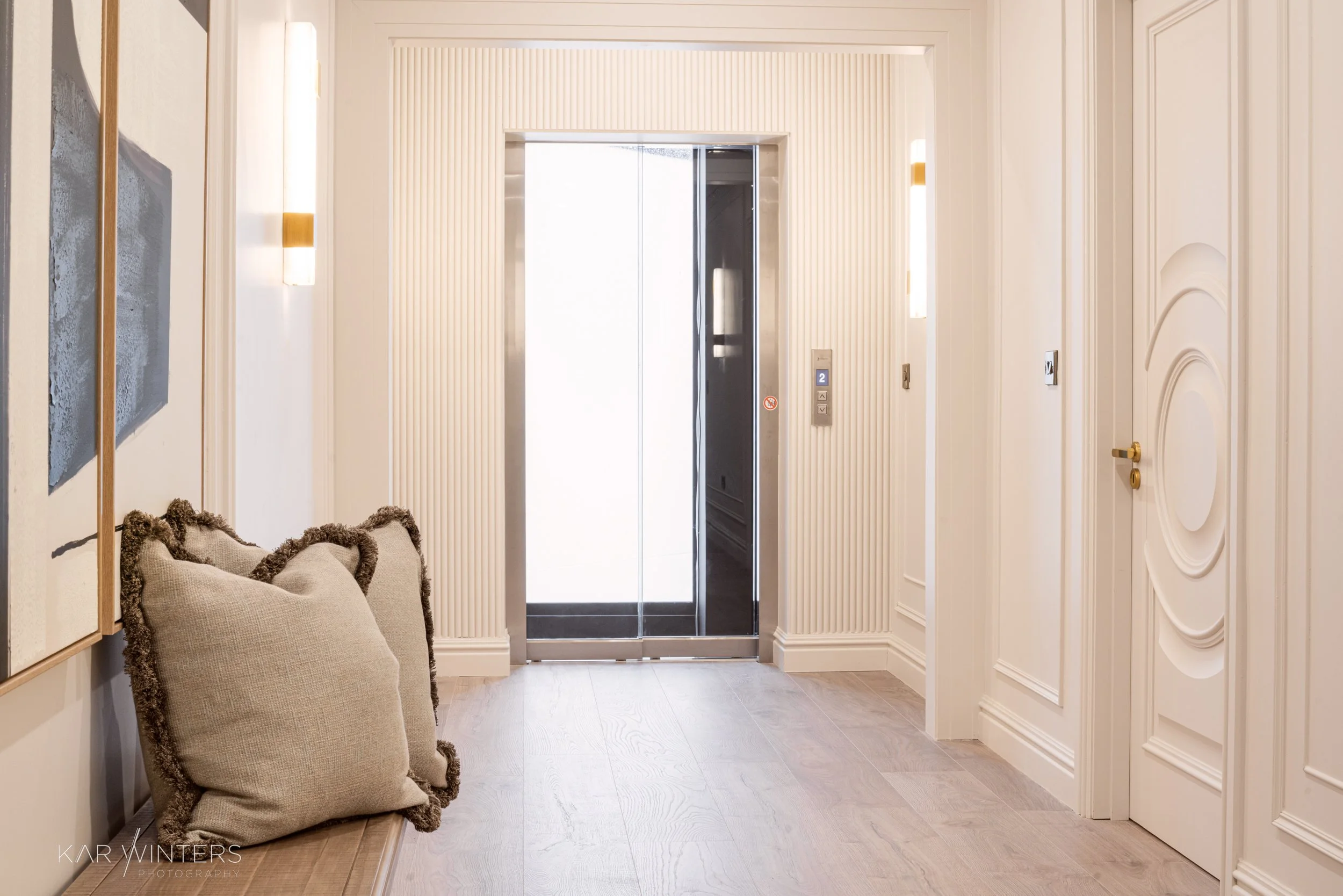 Interior view of an elevator lobby with a closed elevator door, beige walls with decorative paneling, a bench with beige cushions, some artwork on the wall, and subtle lighting.