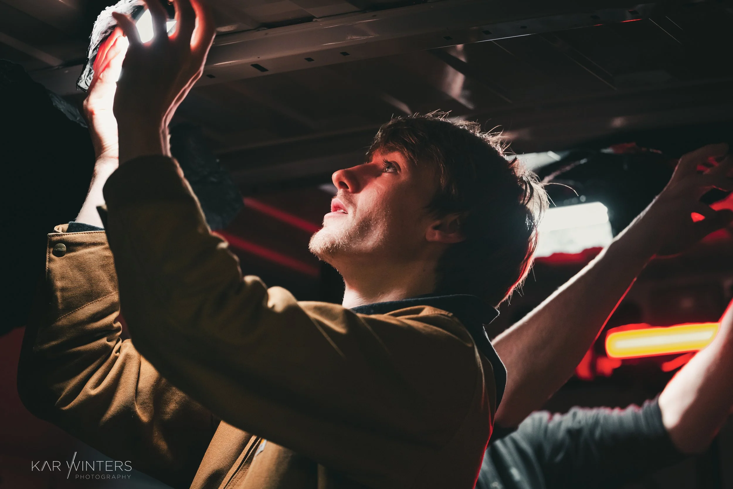 A young man with dark hair and a beard reaching upward with both hands, looking to the side in a dimly lit environment with red and white neon lights.