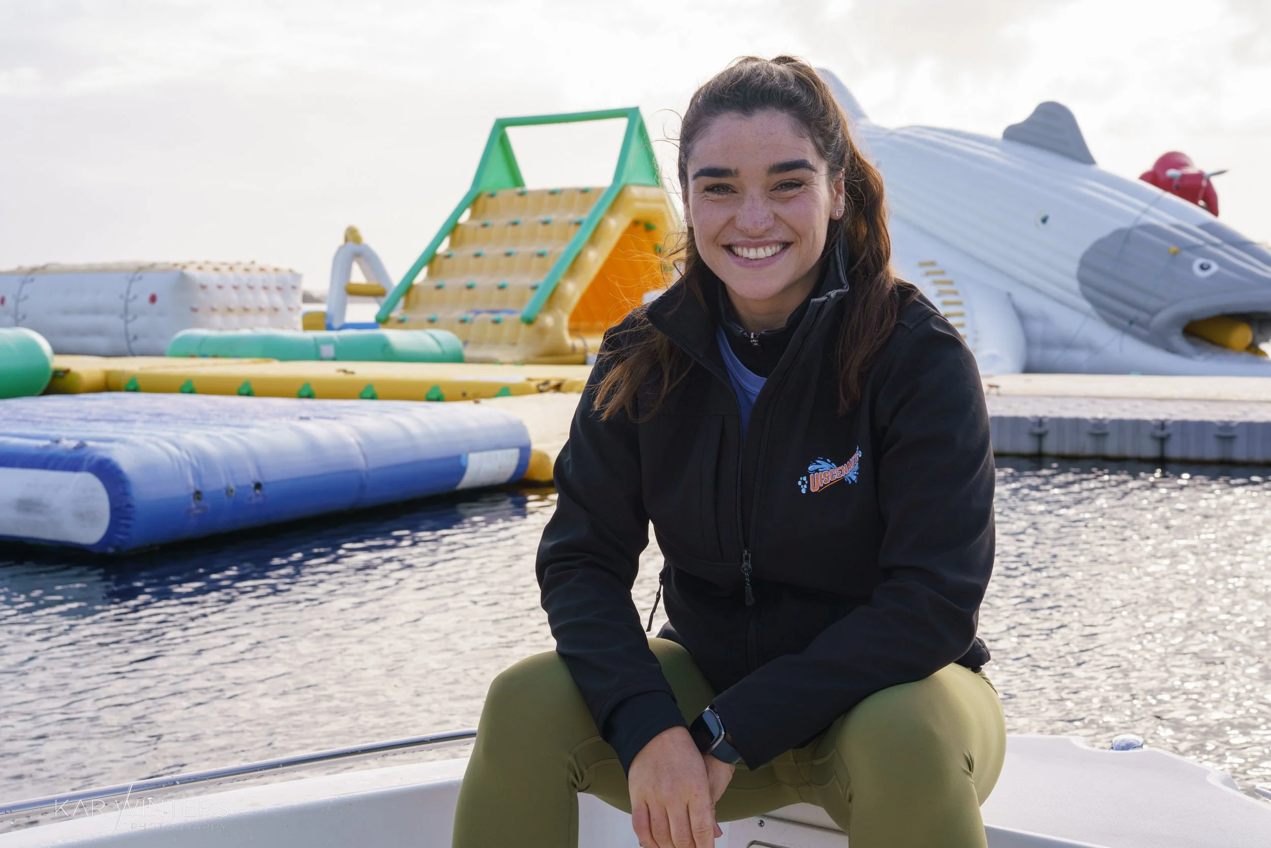 Young woman smiling, sitting on a boat at a floating water park with inflatable slides and obstacle courses behind her.