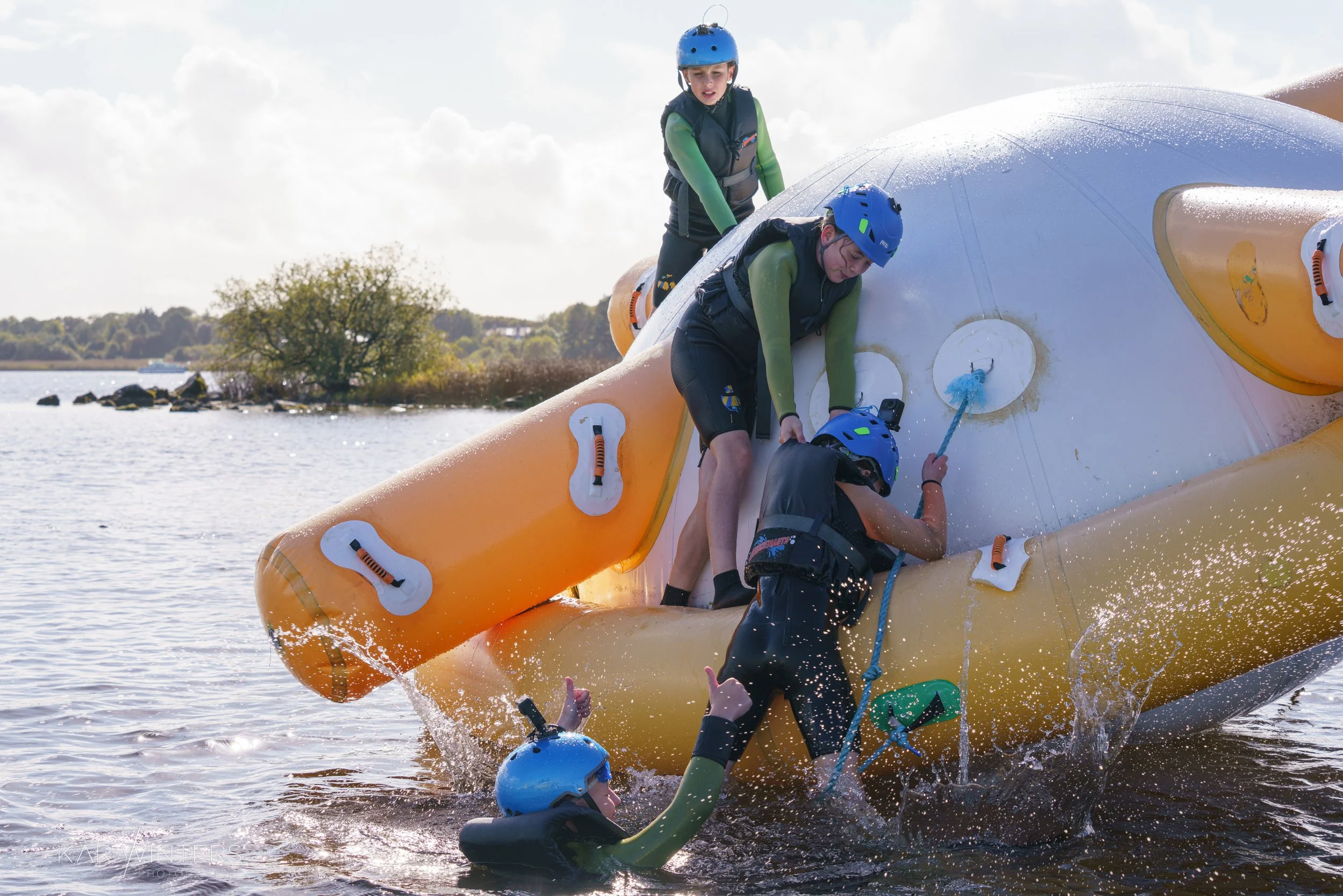Four people wearing helmets and life vests assist a person climbing onto an inflatable watercraft in a lake, with trees and a cloudy sky in the background.