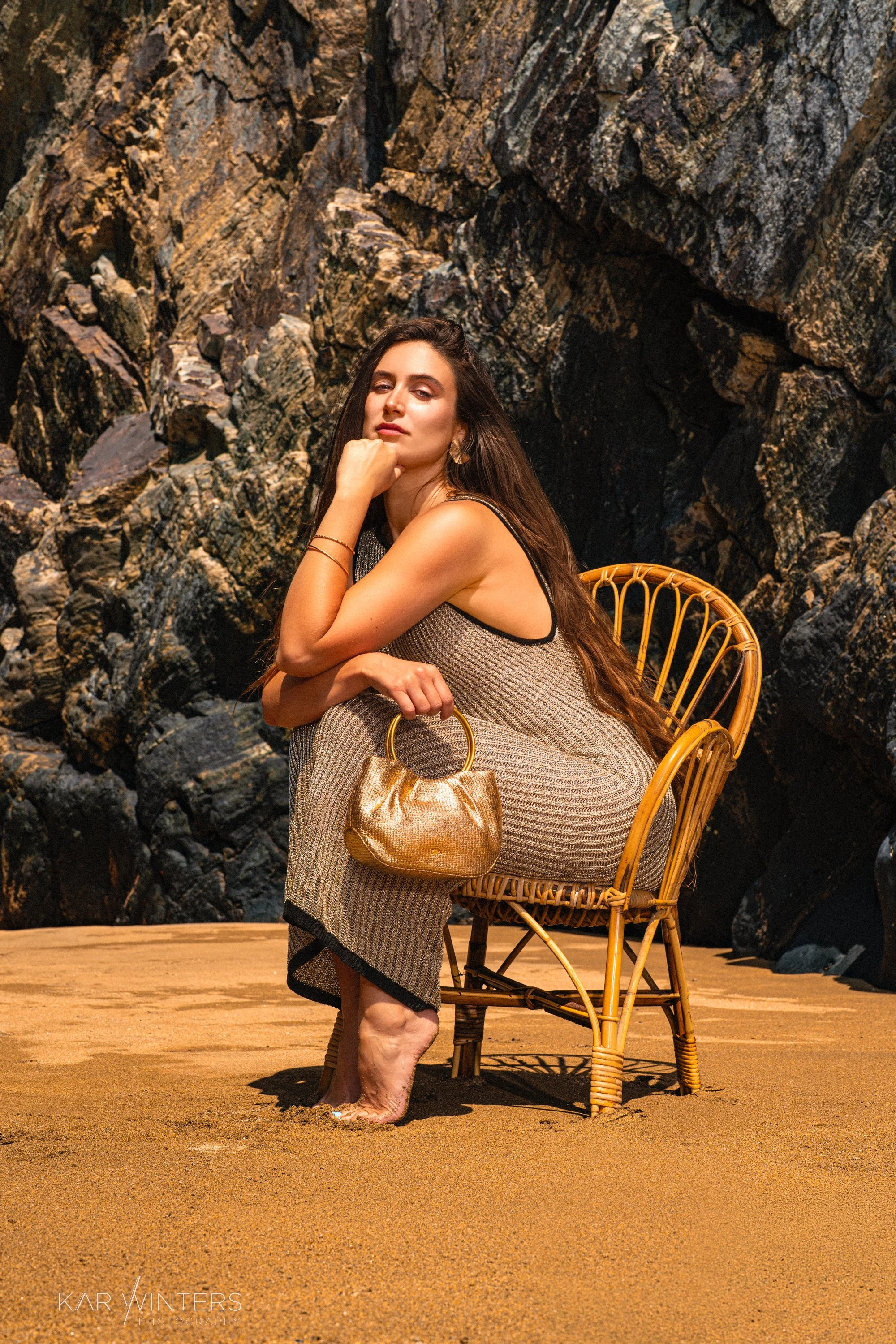 A woman sitting on a wicker chair on a sandy beach with rocky cliffs in the background. She has long dark hair, is wearing a sleeveless dress, jewelry, and is holding a gold handbag.