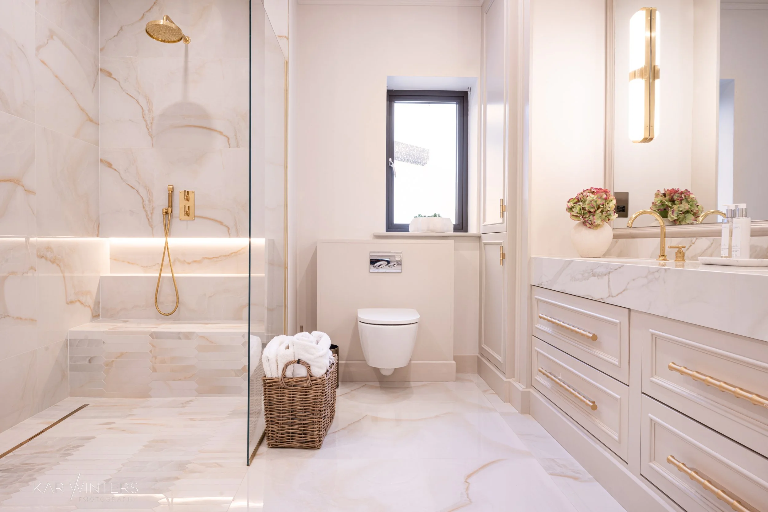 Luxury bathroom with marble floors and walls, a walk-in shower with gold fixtures, a wall-mounted toilet, white cabinetry with gold handles, a window, and decorative flowers.