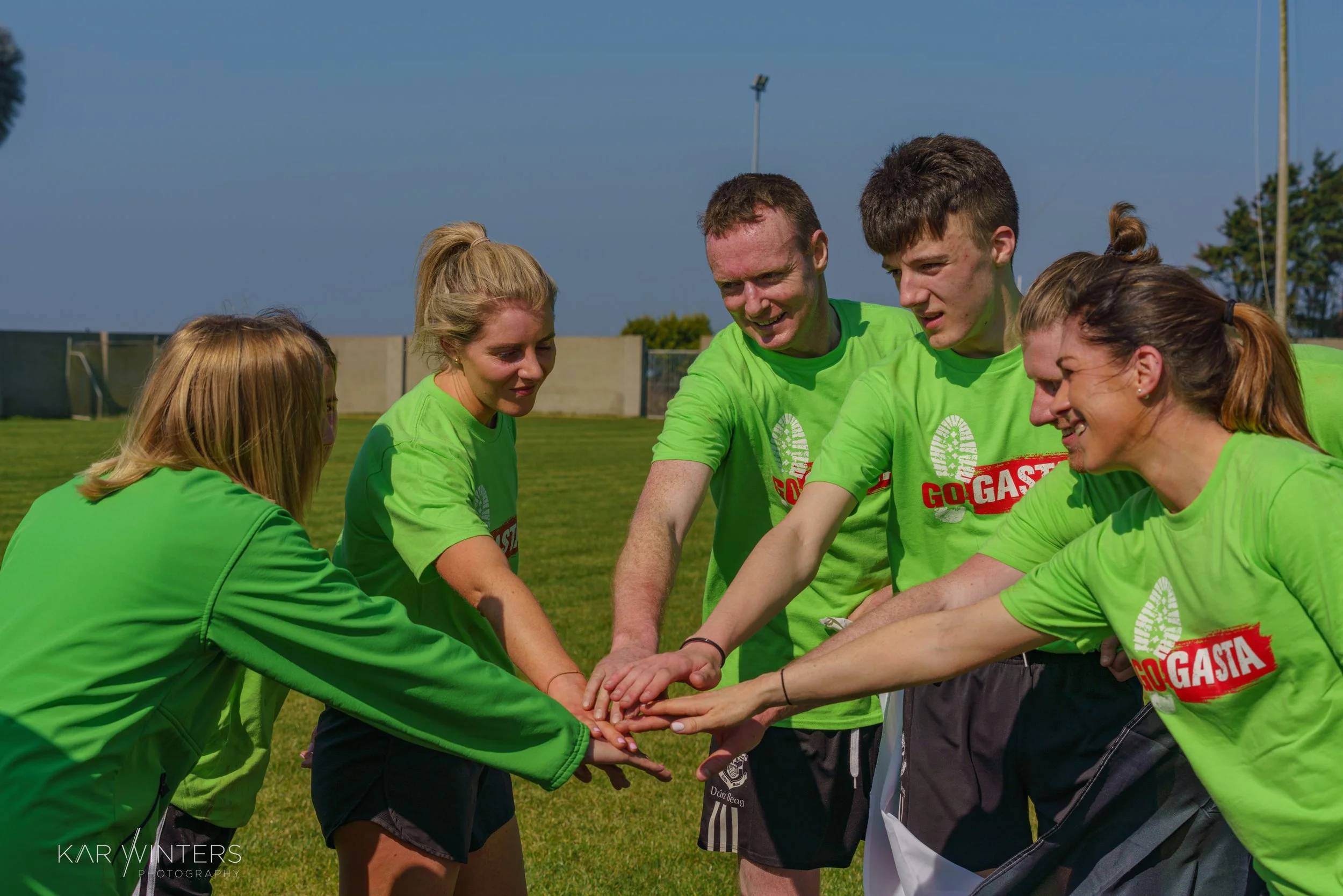 Group of six young adults in bright green shirts stacking hands together on a soccer field.