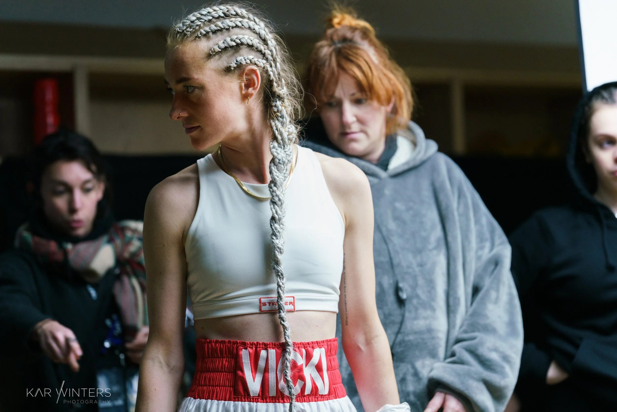 A female boxer with long braided hair and a white sports bra stands in a gym, with three women in the background looking down or away.