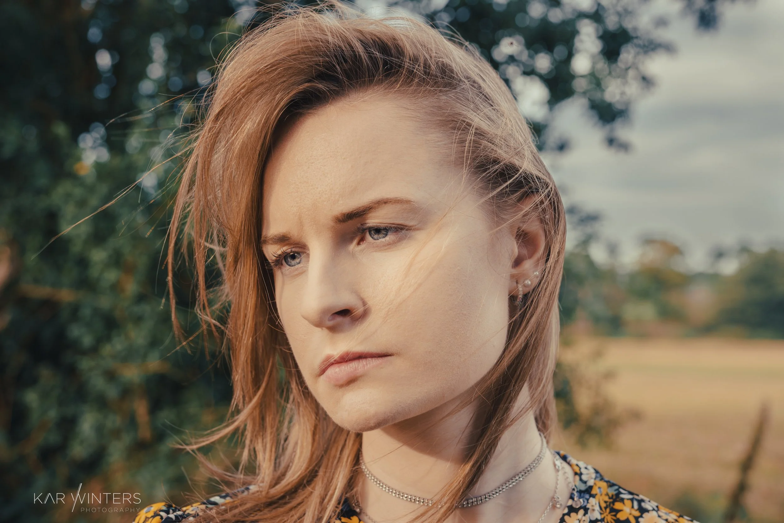A young woman with red hair looking contemplative outdoors with trees and open field in the background.