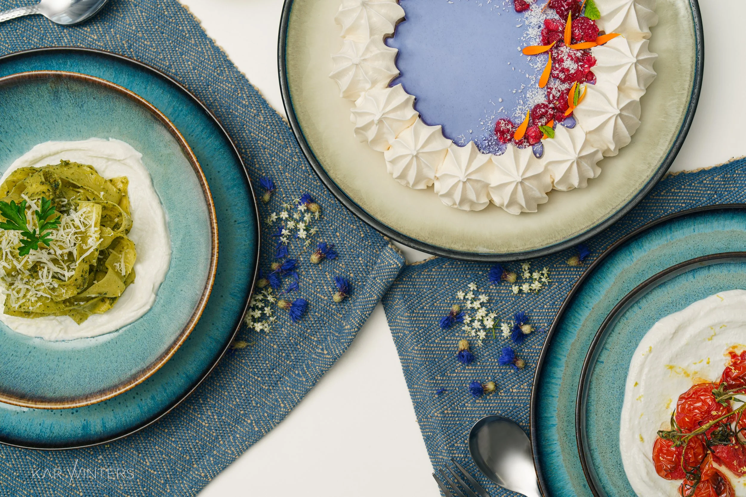 Three dishes served on blue-green plates. The top dish is a dessert cake with white frosting and red fruit toppings, decorated with flower petals. The left dish is pasta on a bed of white sauce, garnished with green herbs and grated cheese. The right