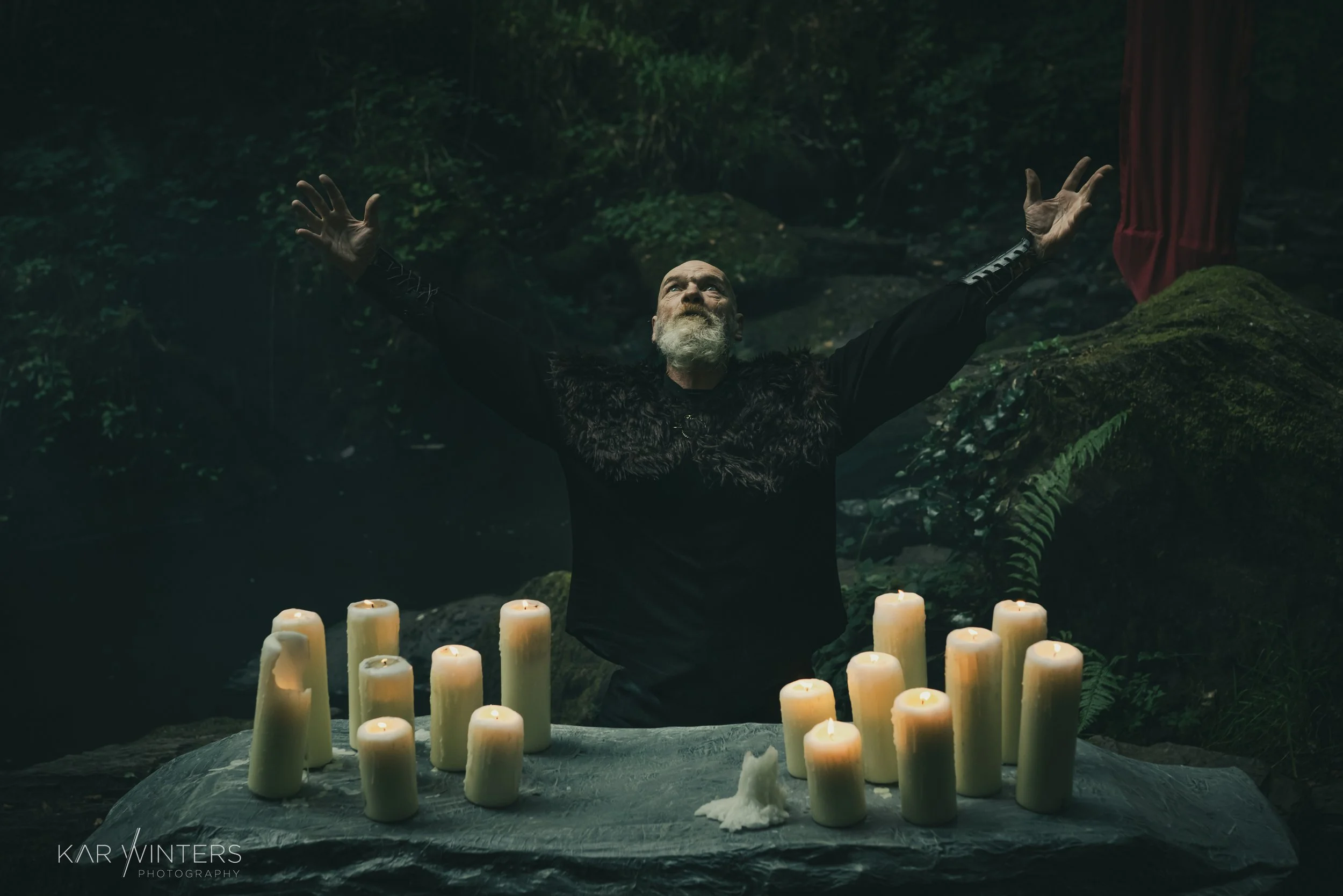 A bearded man with a black fur and leather robe standing with arms outstretched behind a table with lit candles in a dark, forested setting.