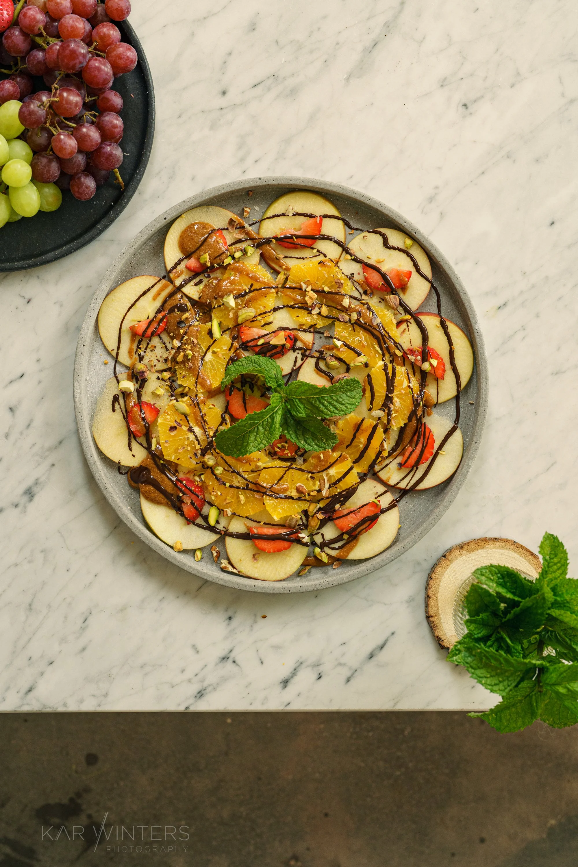 Sliced apples topped with chocolate drizzle, chopped nuts, and fruit slices on a gray plate, with fresh grapes on a black plate nearby on a white marble surface.
