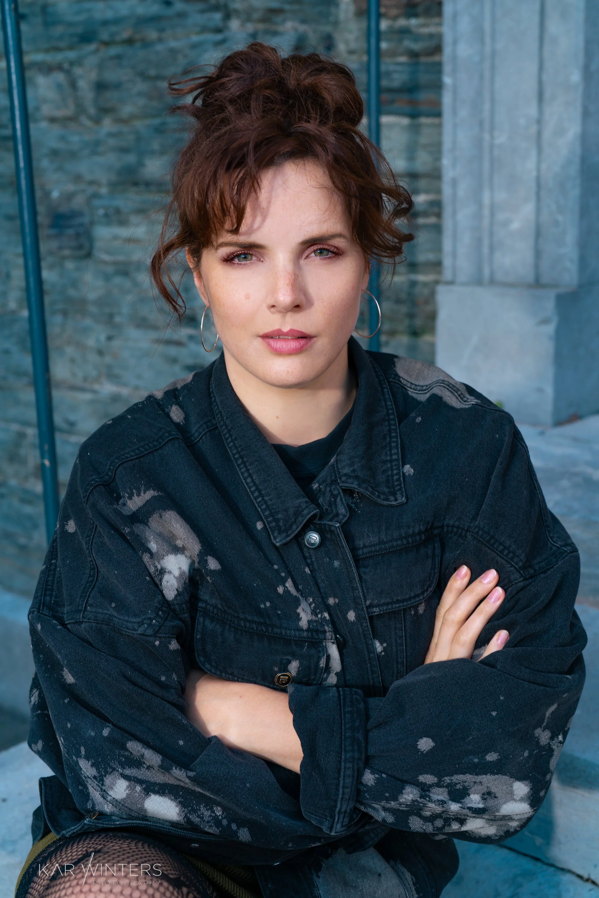 A young woman with short curly red hair, wearing hoop earrings and a black tie-dye denim jacket, sitting outdoors with crossed arms, looking into the camera against a background of stone steps and blue railings.