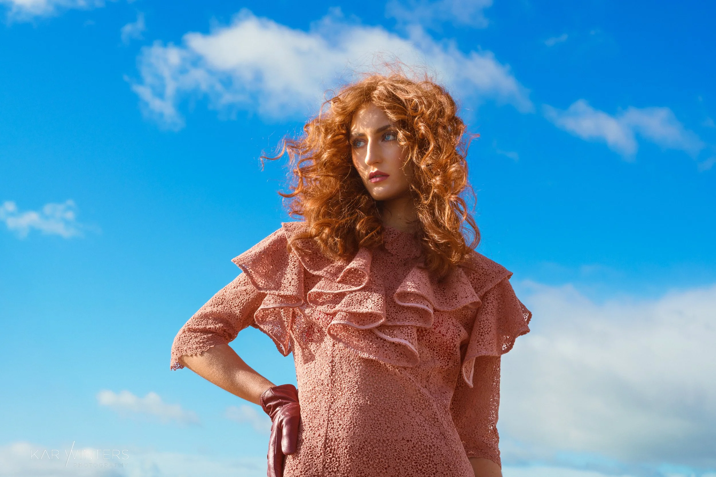 A woman with curly red hair wearing a pink lace dress with ruffles on the shoulders, standing outdoors against a blue sky with some clouds.