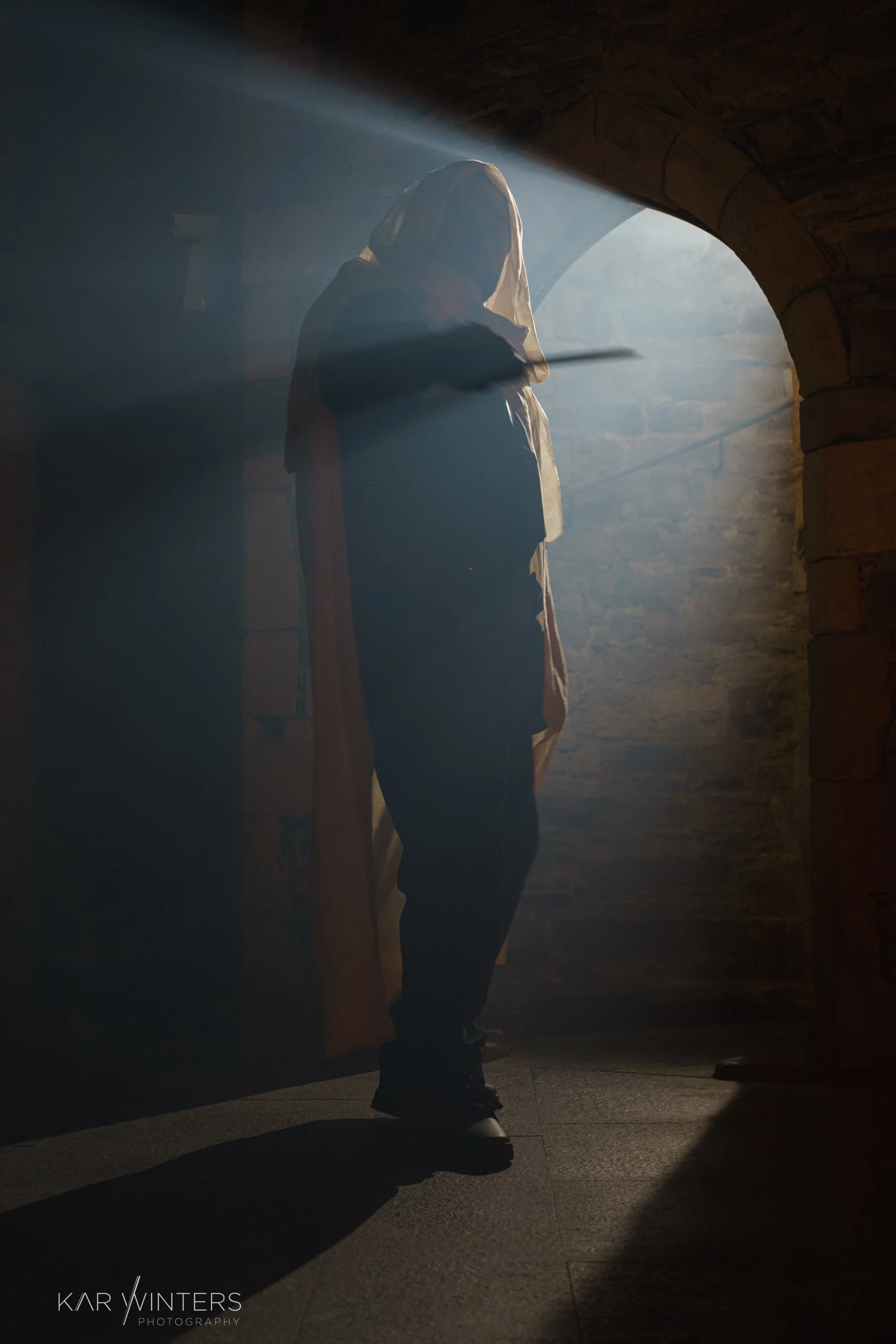A person wearing a hooded cloak standing in a dimly lit stone corridor with sunlight streaming through an archway, creating a dramatic shadow.
