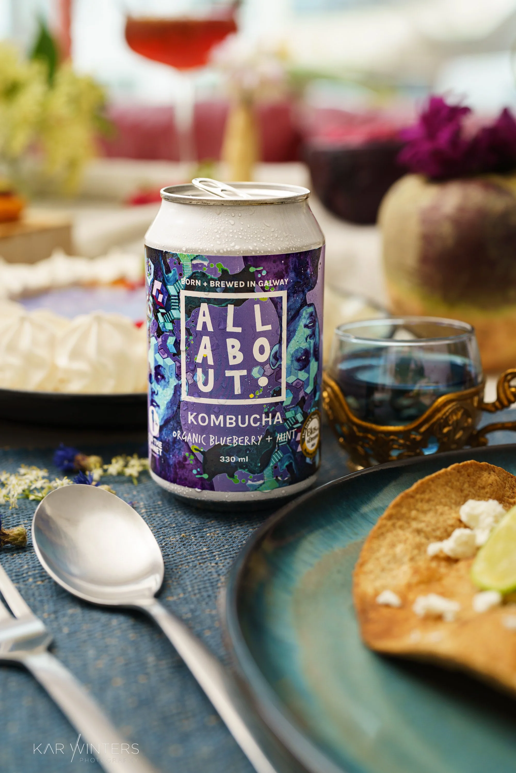 A can of All About Kombucha with organic blueberry and mint flavor on a table, surrounded by a spoon, a plate with food, and a glass of water, with a blurred background of a table setting and decor.