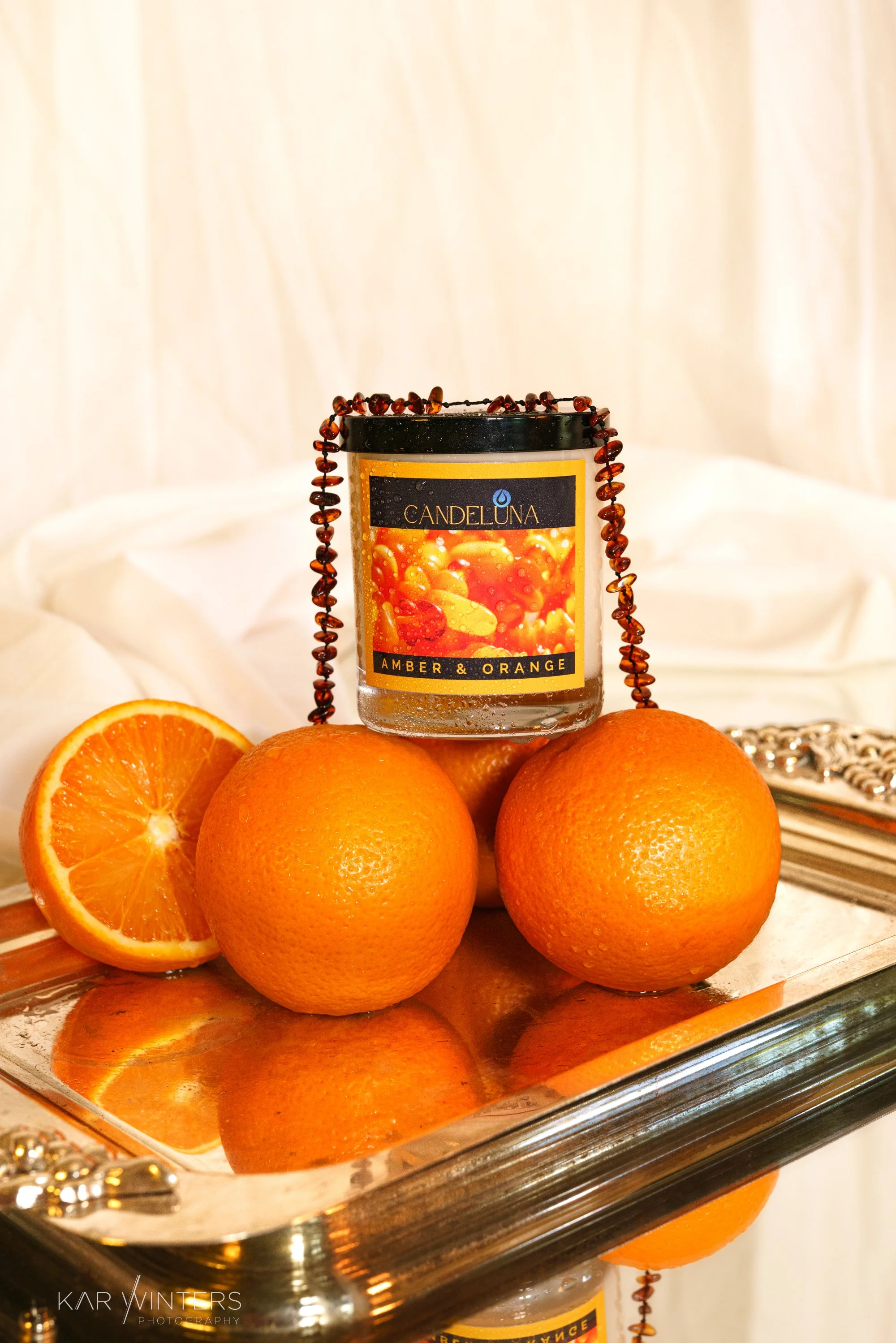 A silver tray holding three whole oranges and one orange half, with a candle labeled 'Candeluna Amber & Orange' placed behind the oranges, and a beaded necklace draped around the candle.