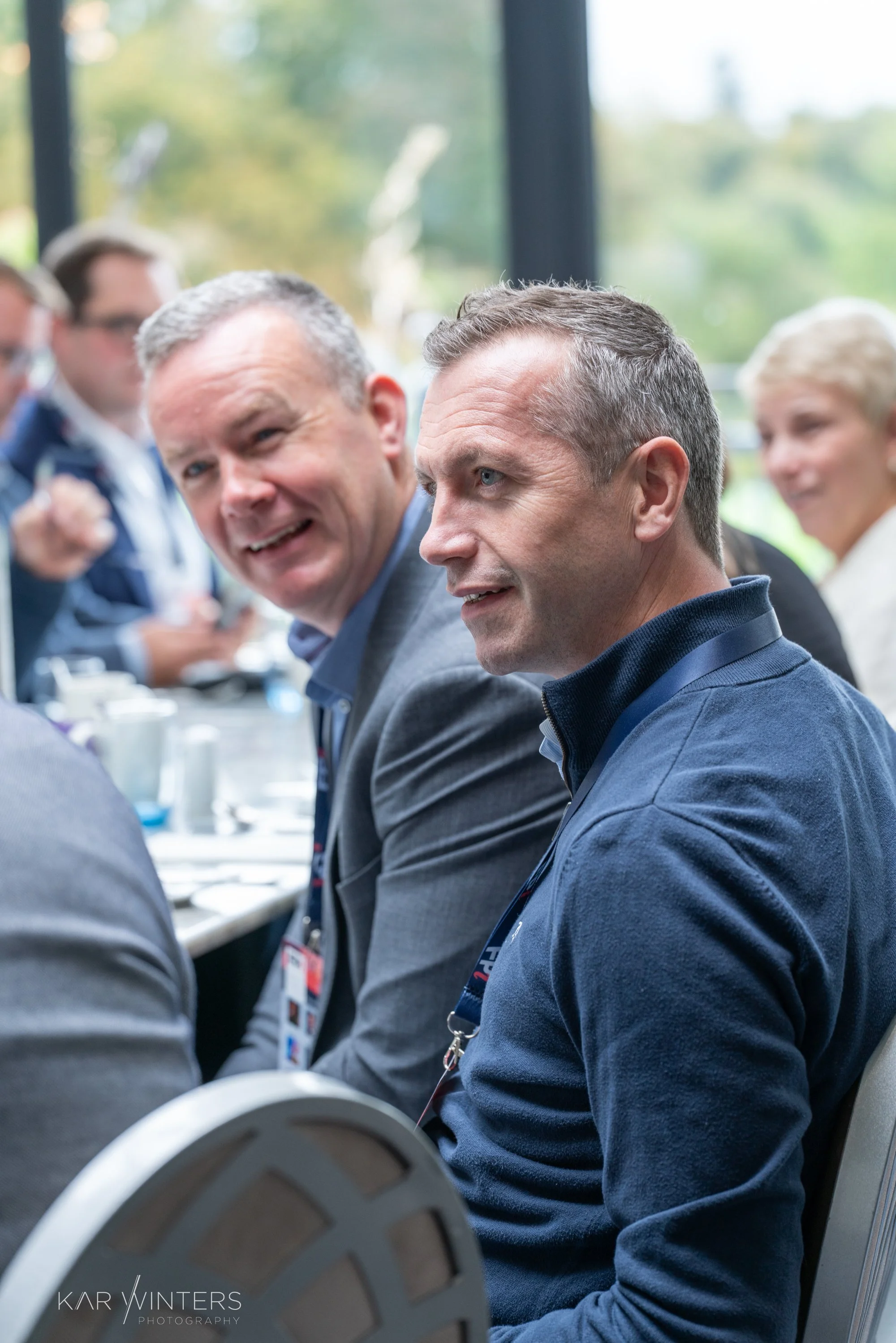 A group of people sitting at a conference table, engaging in a discussion, with two men prominently in focus in the foreground.