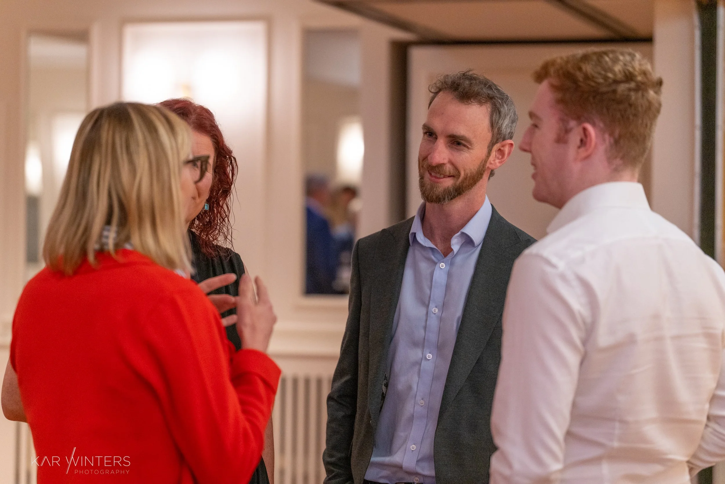 Group of four people engaged in conversation at an indoor social event, smiling and interacting with each other.