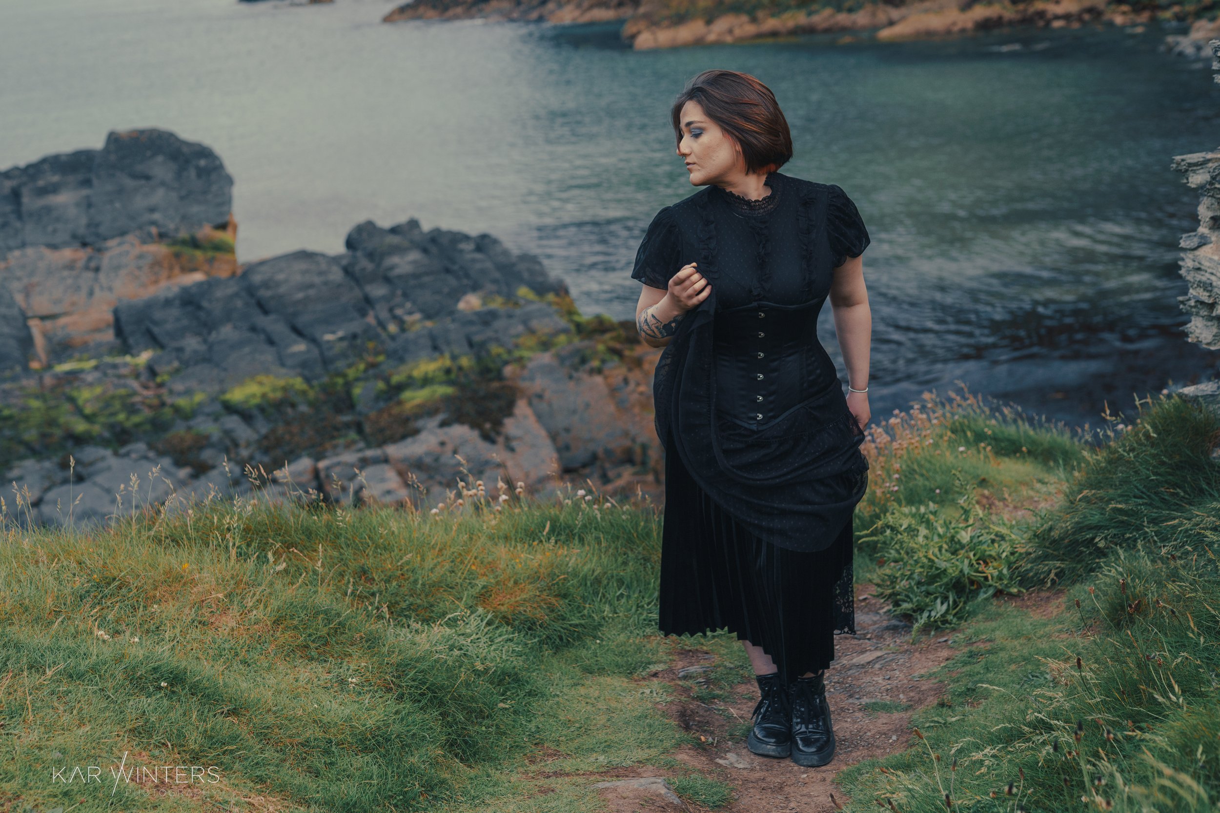 A woman with dark hair in a bob cut, wearing a long black dress and black boots, standing on a grassy path near a rocky shoreline, looking to the side with her hand on her chest, by the ocean.