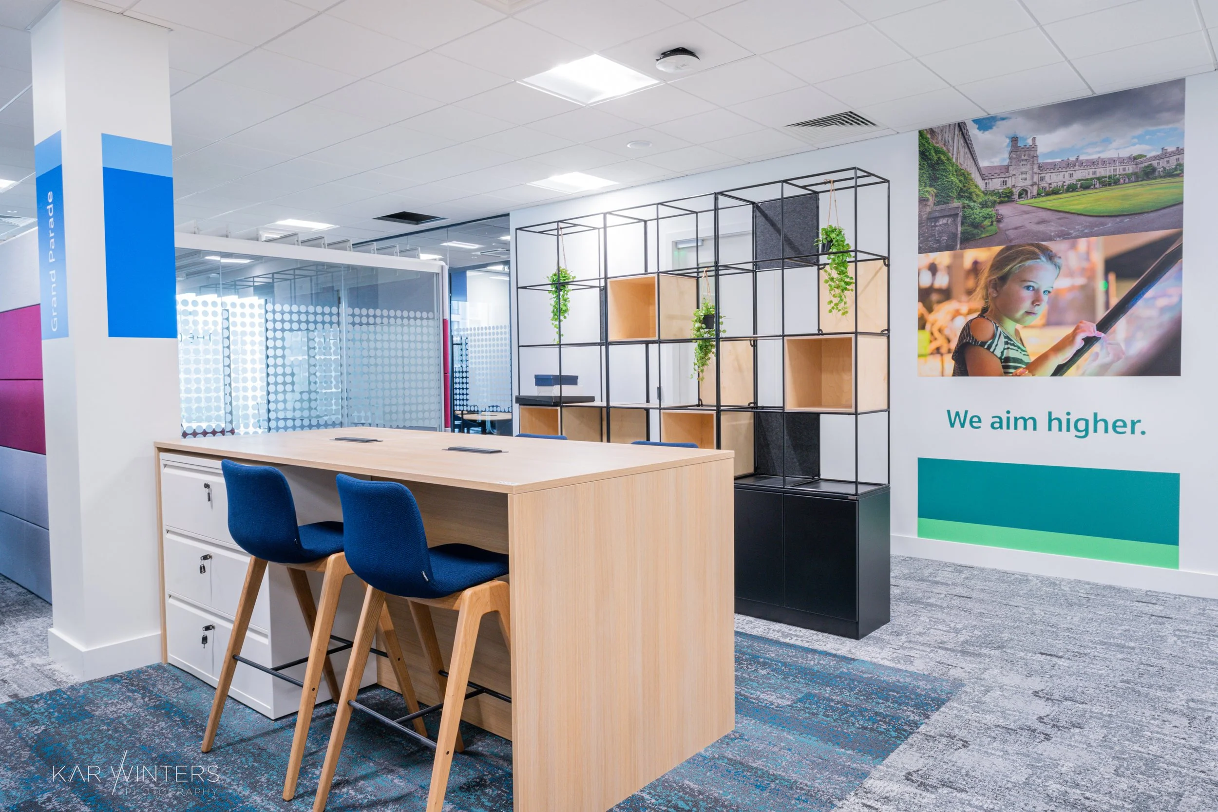 Modern office space with a wooden table, two blue chairs, and a black shelving unit with plants. There is a large wall poster of a young girl using a touchscreen device and the phrase "We aim higher." The office has glass partitions, white walls, gra