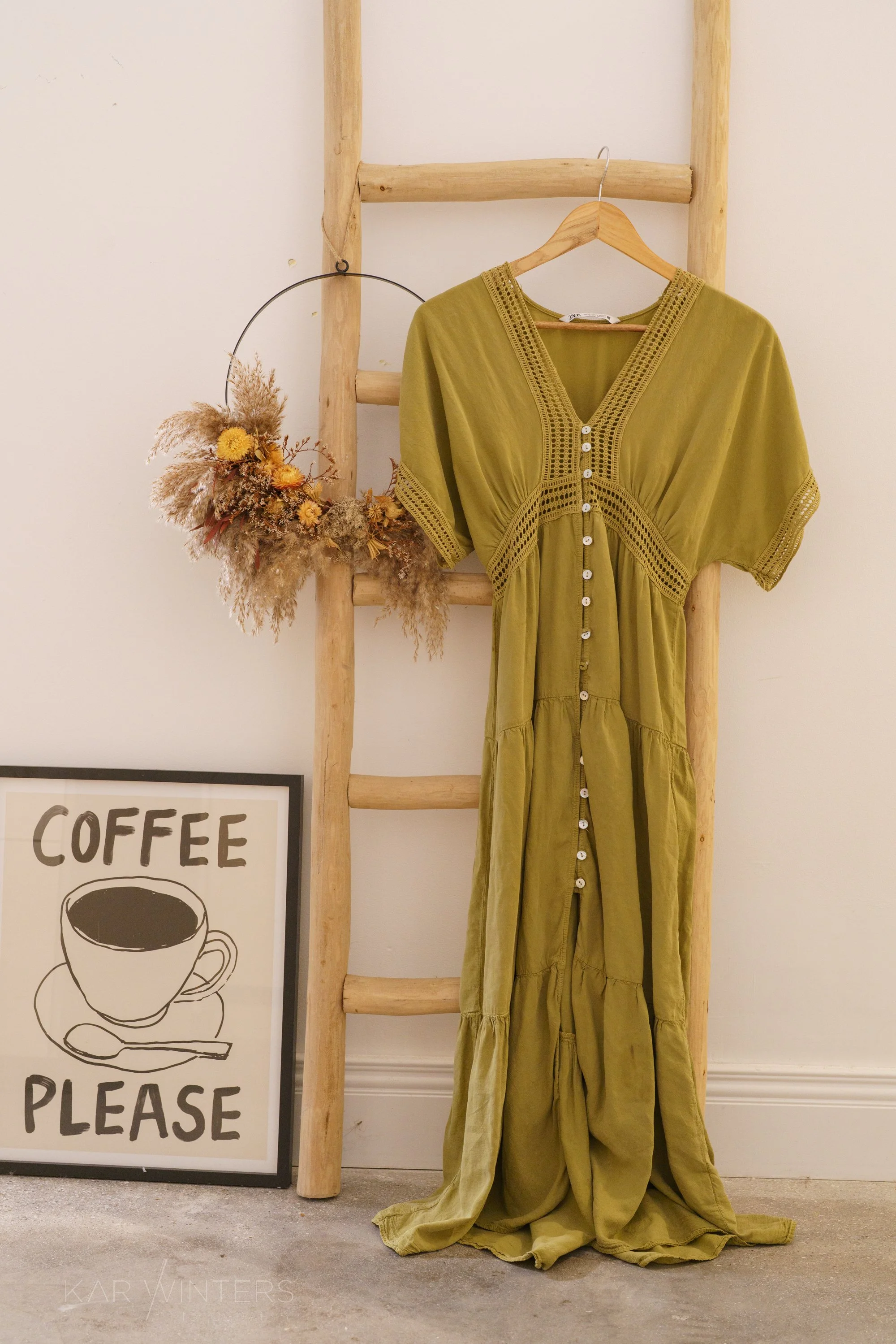 A green dress hanging on a wooden ladder with a floral arrangement on the side. A coffee sign is partially visible in the lower left corner.