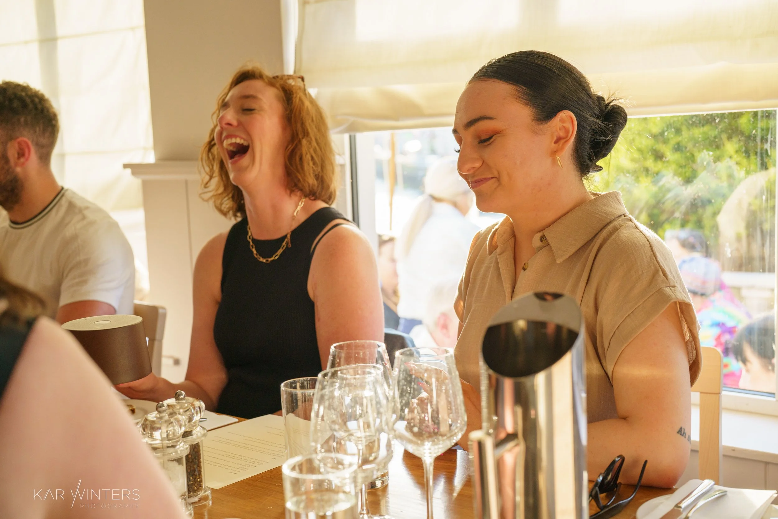 Two women sitting at a restaurant table, smiling and laughing. The woman on the left has red hair and is wearing a black sleeveless top, while the woman on the right has dark hair tied back and is wearing a beige shirt. There are glasses and a stainl