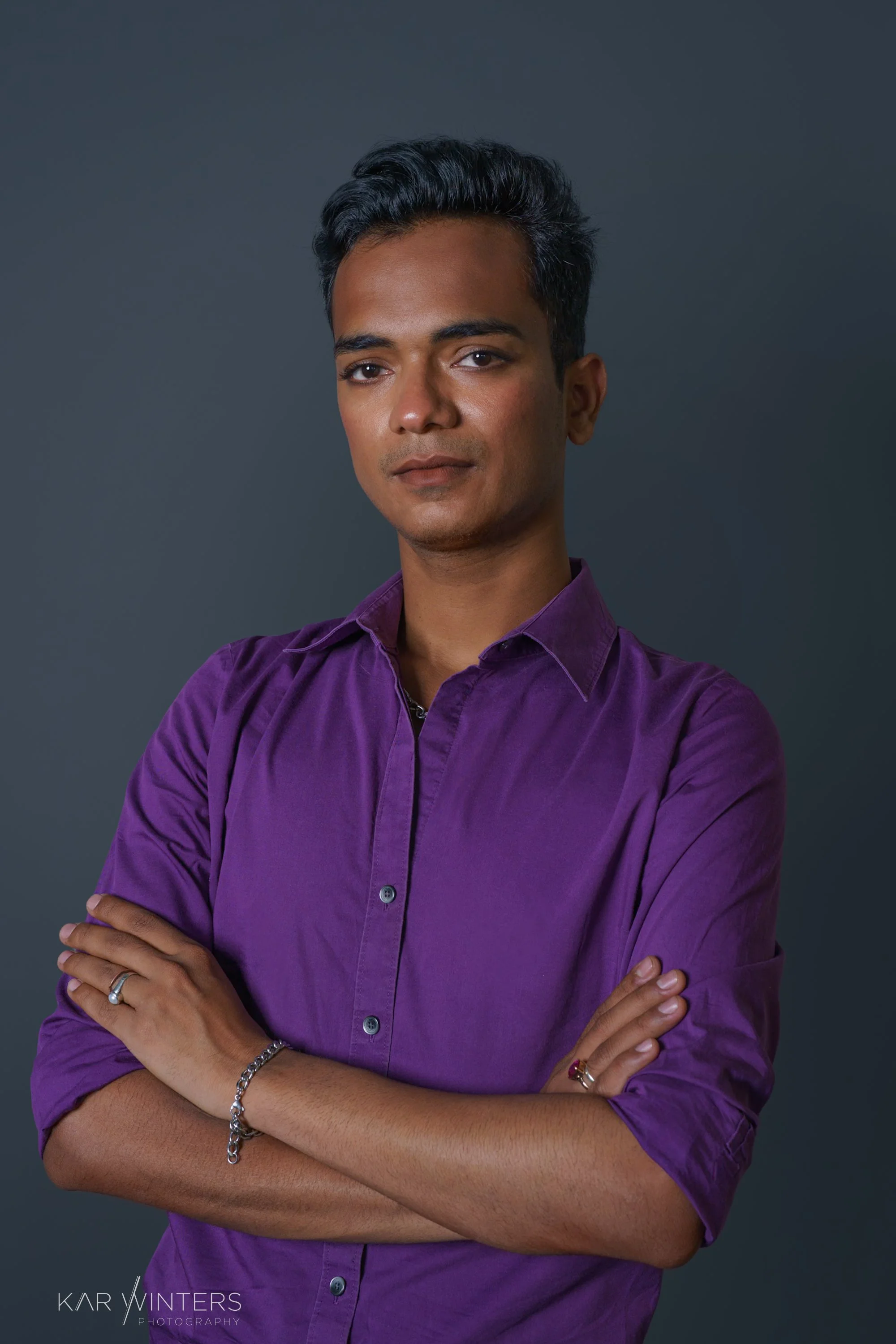 A young man with dark hair, wearing a purple button-up shirt, standing with arms crossed against a dark background.