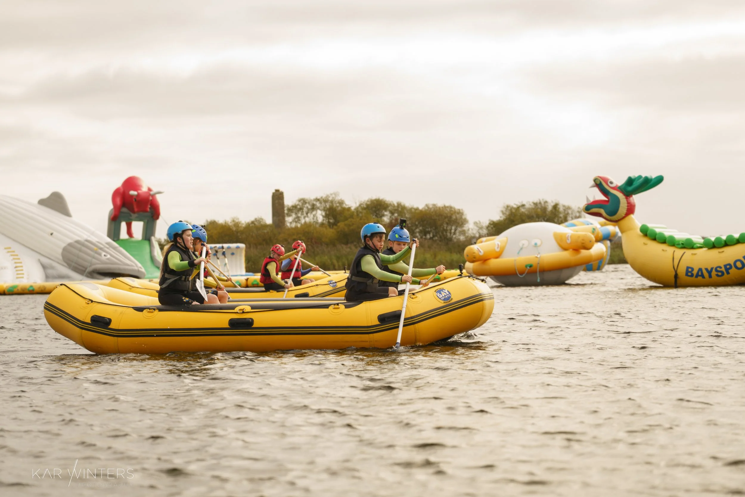 Children wearing life jackets and helmets paddling in a yellow inflatable raft on a body of water, with colorful inflatable animal floats in the background.