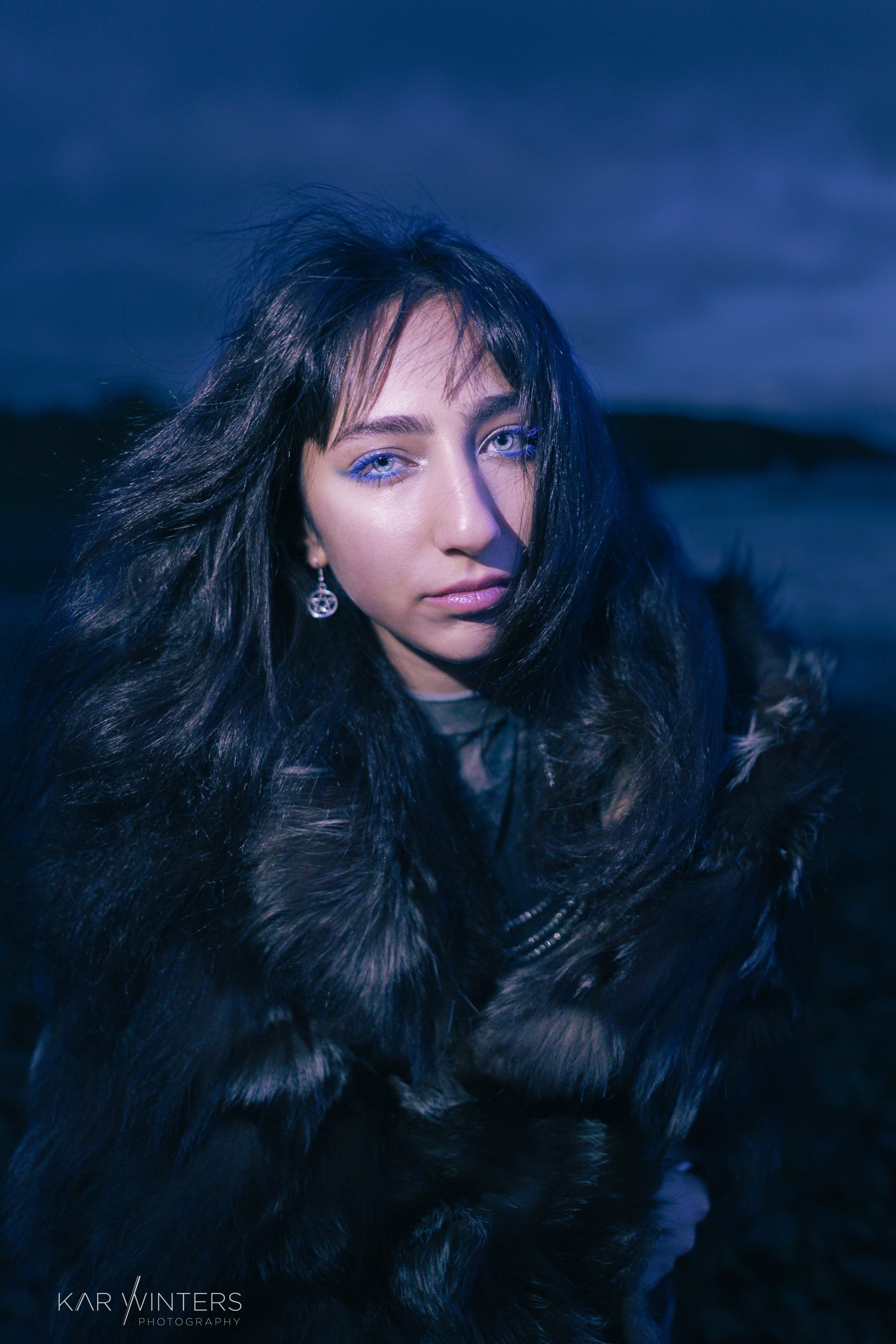 A young woman with blue eyes and long dark hair wearing a fur coat, standing outdoors near a body of water during twilight.