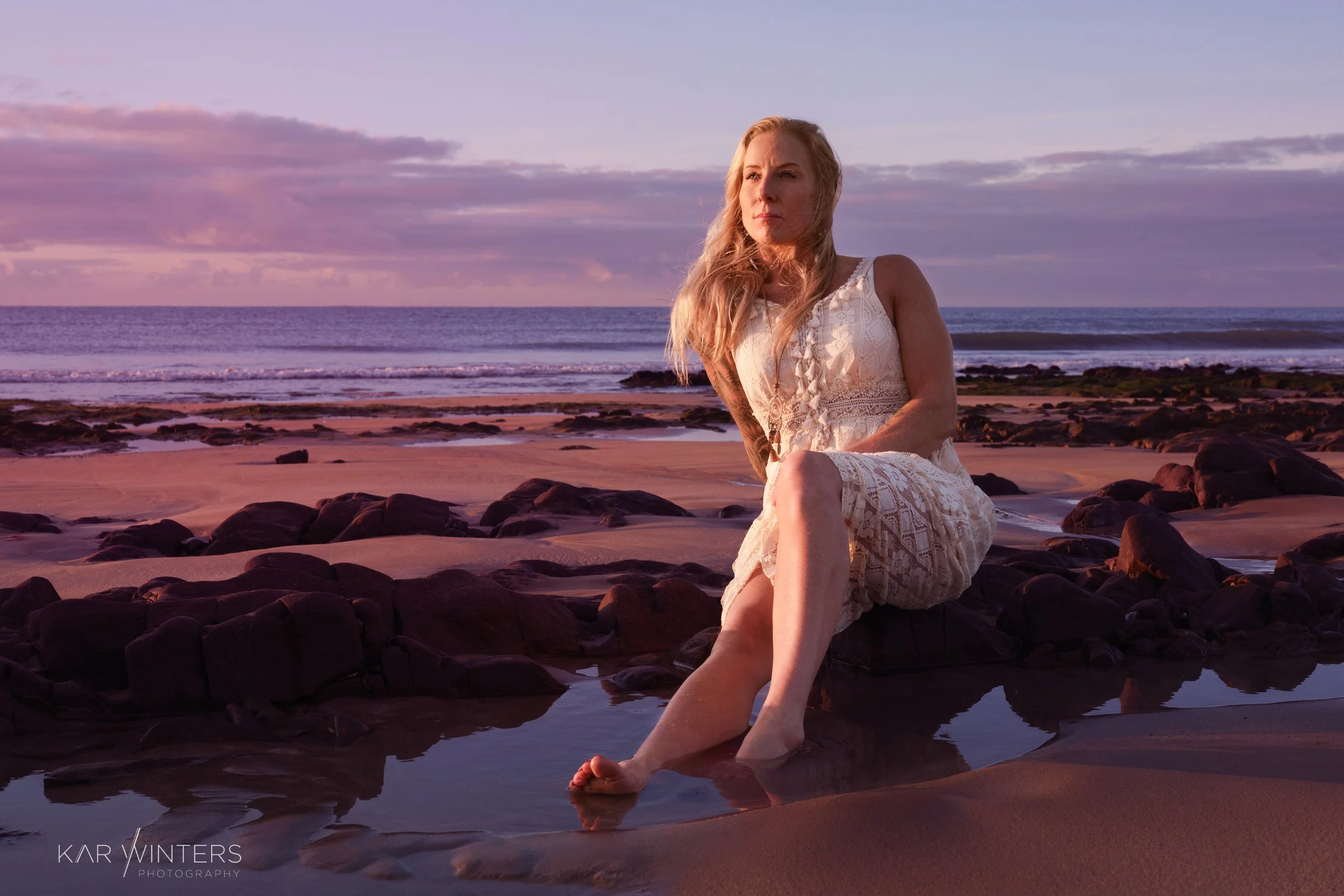 A woman in a white lace dress sitting on rocks at the beach during sunset, with the ocean and sky in the background.