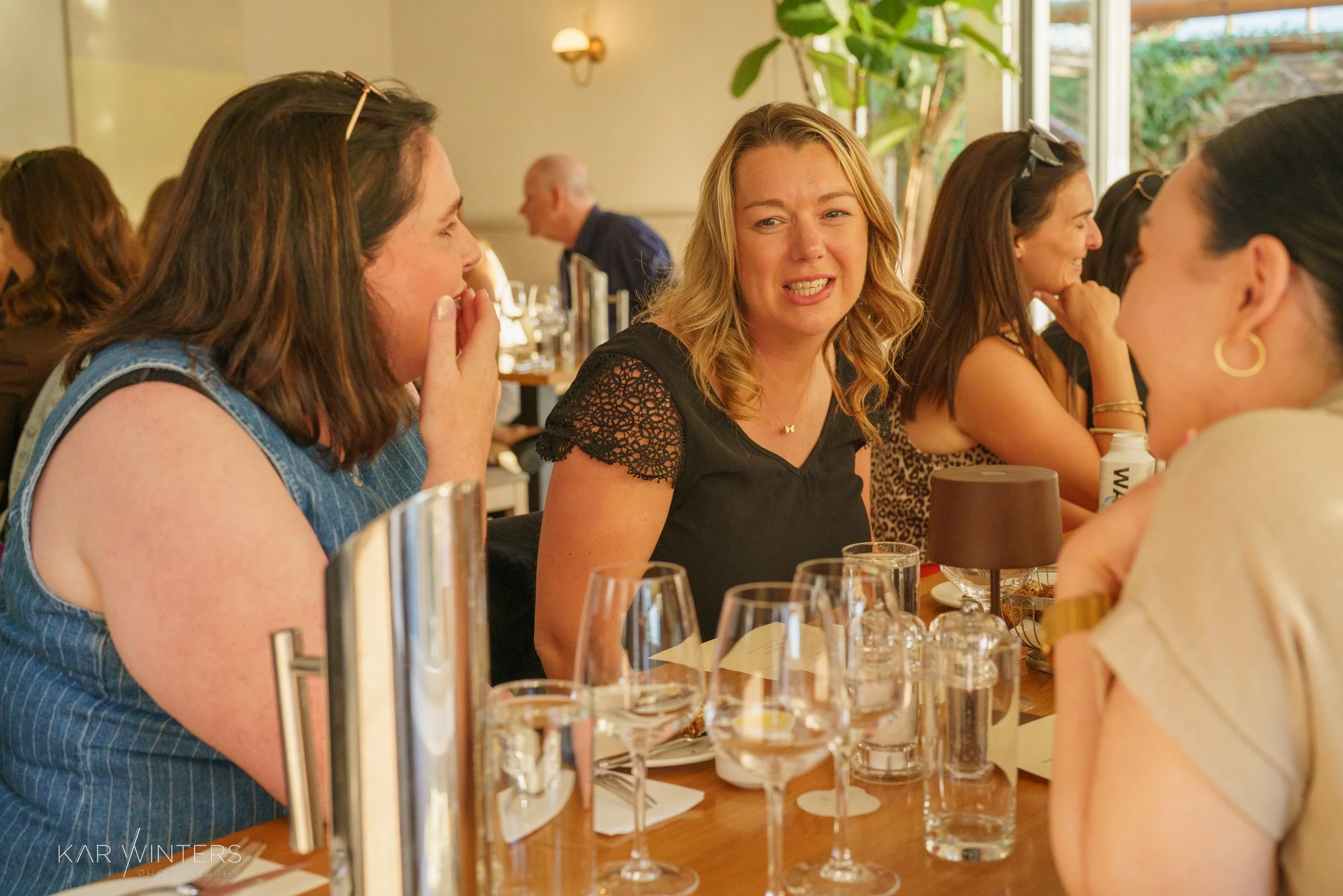 Women sitting at a social gathering, engaging in conversation, with glasses and drinks on the table.