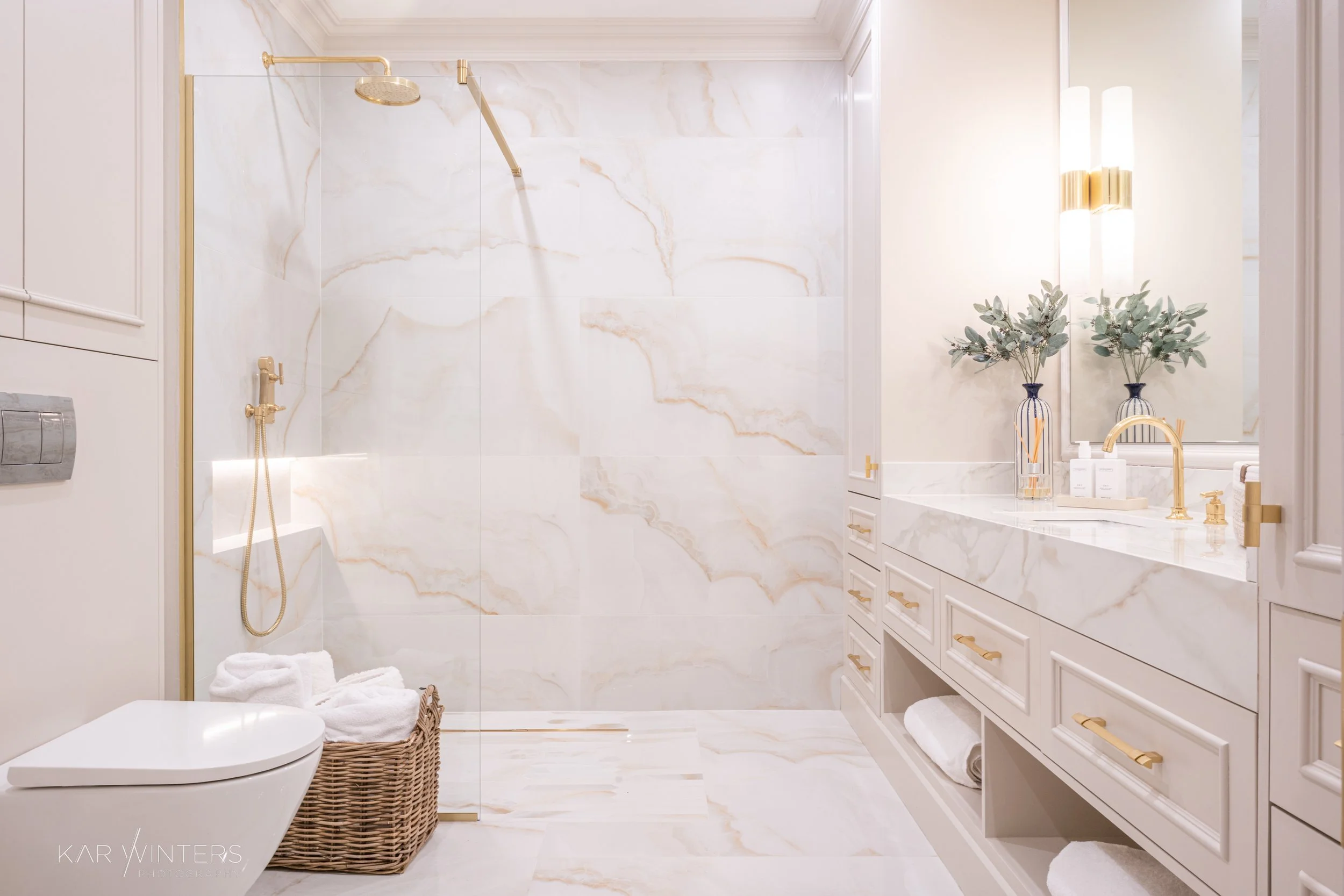 Luxurious bathroom with a walk-in shower enclosed with clear glass, featuring a gold showerhead and fixtures, a white toilet, a woven basket with towels, a white marble vanity with gold handles, a large mirror above the vanity, and decorative vases w