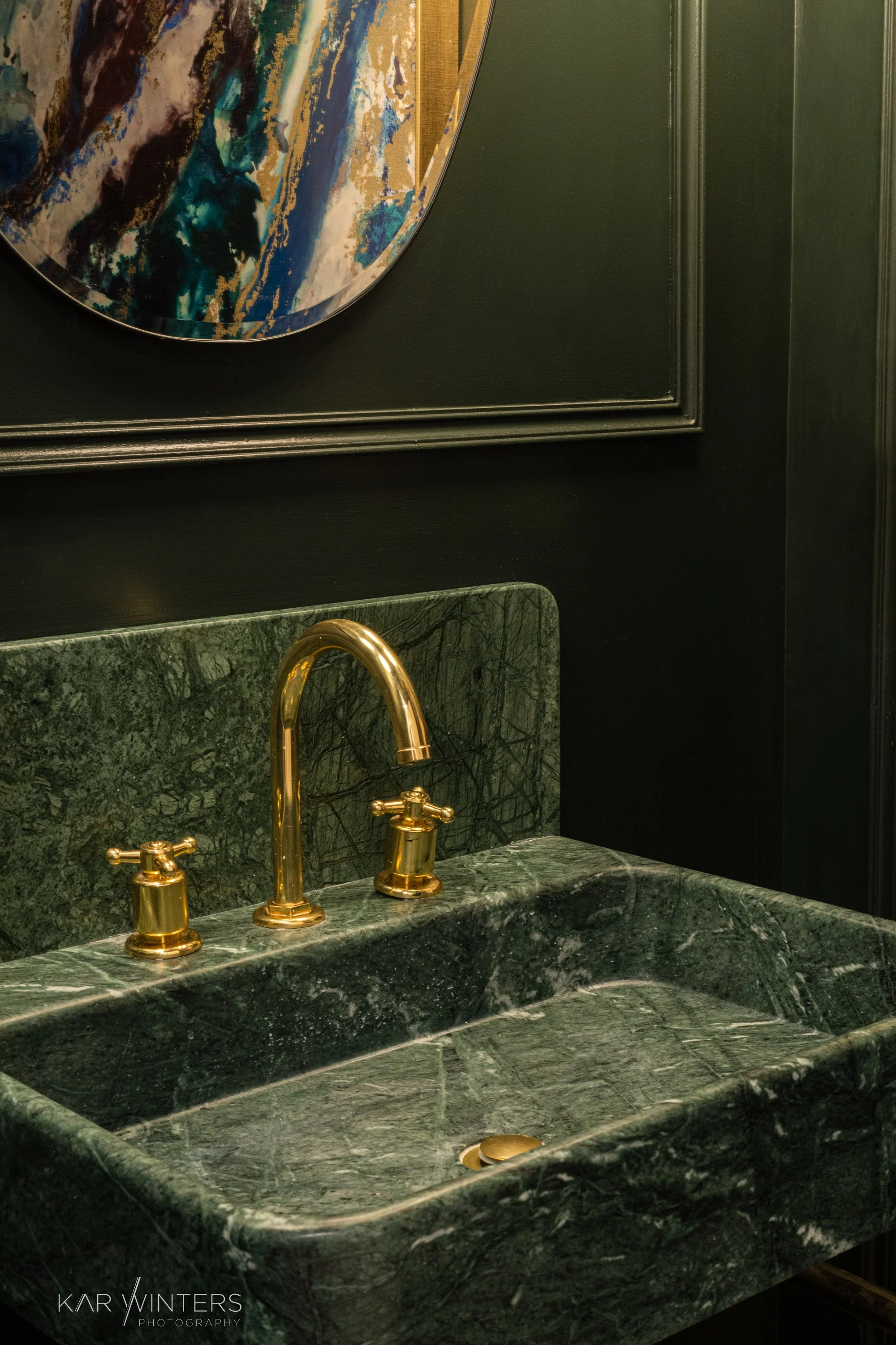 A green marble sink with gold fixtures, including a faucet and two handles, set against a dark wall with a colorful abstract painting above.