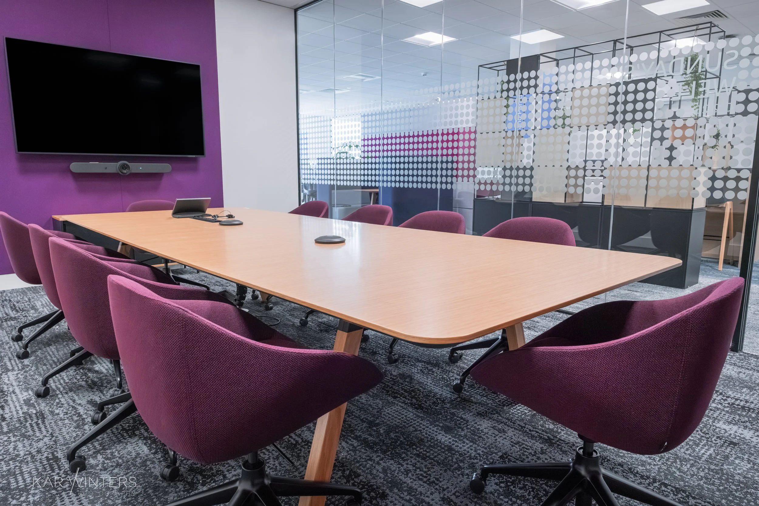 Empty conference room with a long wooden table, nine maroon office chairs, a large wall-mounted black screen with a video conference camera underneath, a laptop with a mouse on the table, a glass partition with dotted white patterns, and a purple acc
