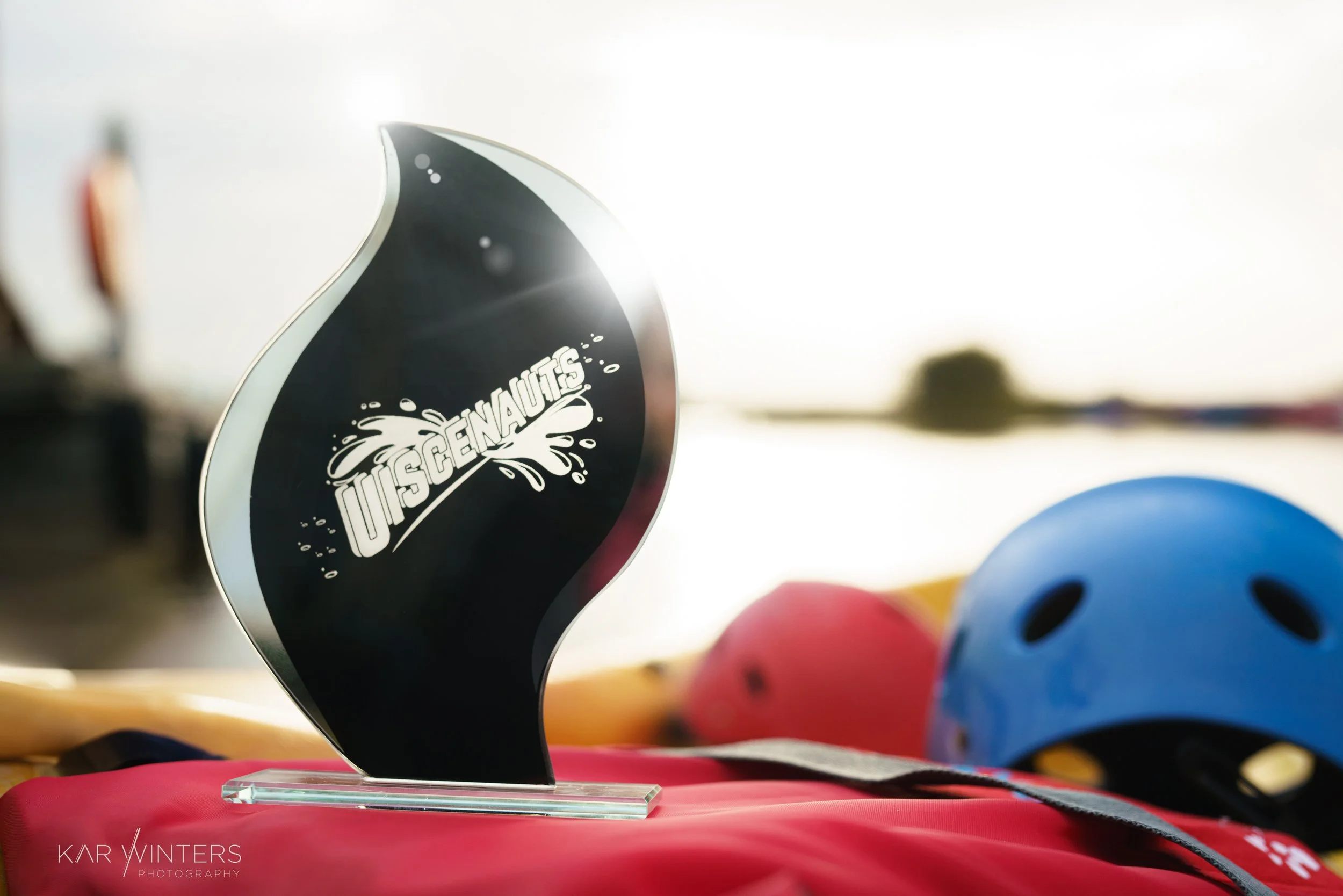 A black and clear glass award shaped like a flame with the word "USCENNAU" written in white, placed on a red cloth with helmets in the background.