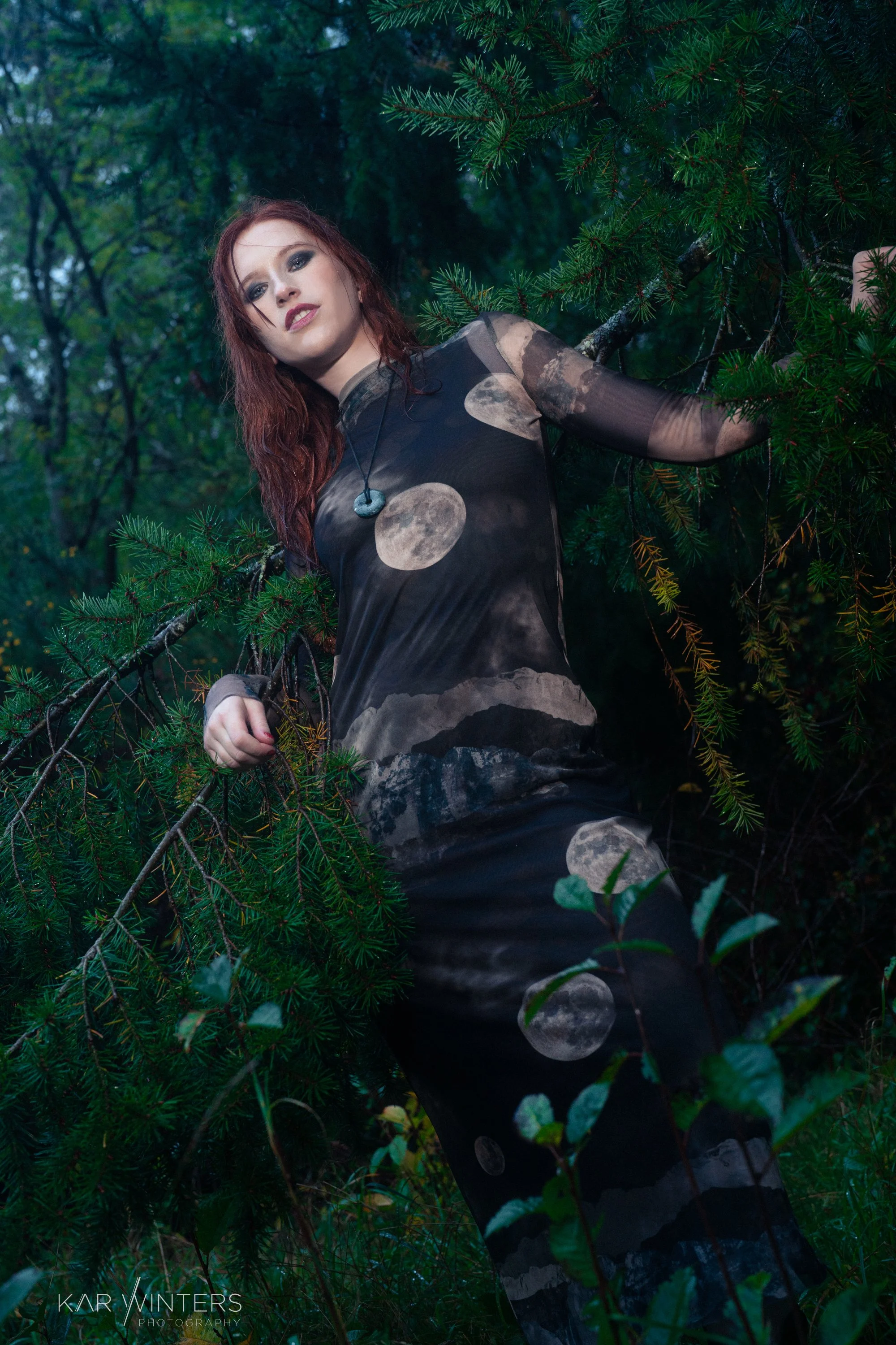 A woman with red hair, wearing a black dress with moon phase patterns, stands among dense green trees, in a forest setting.