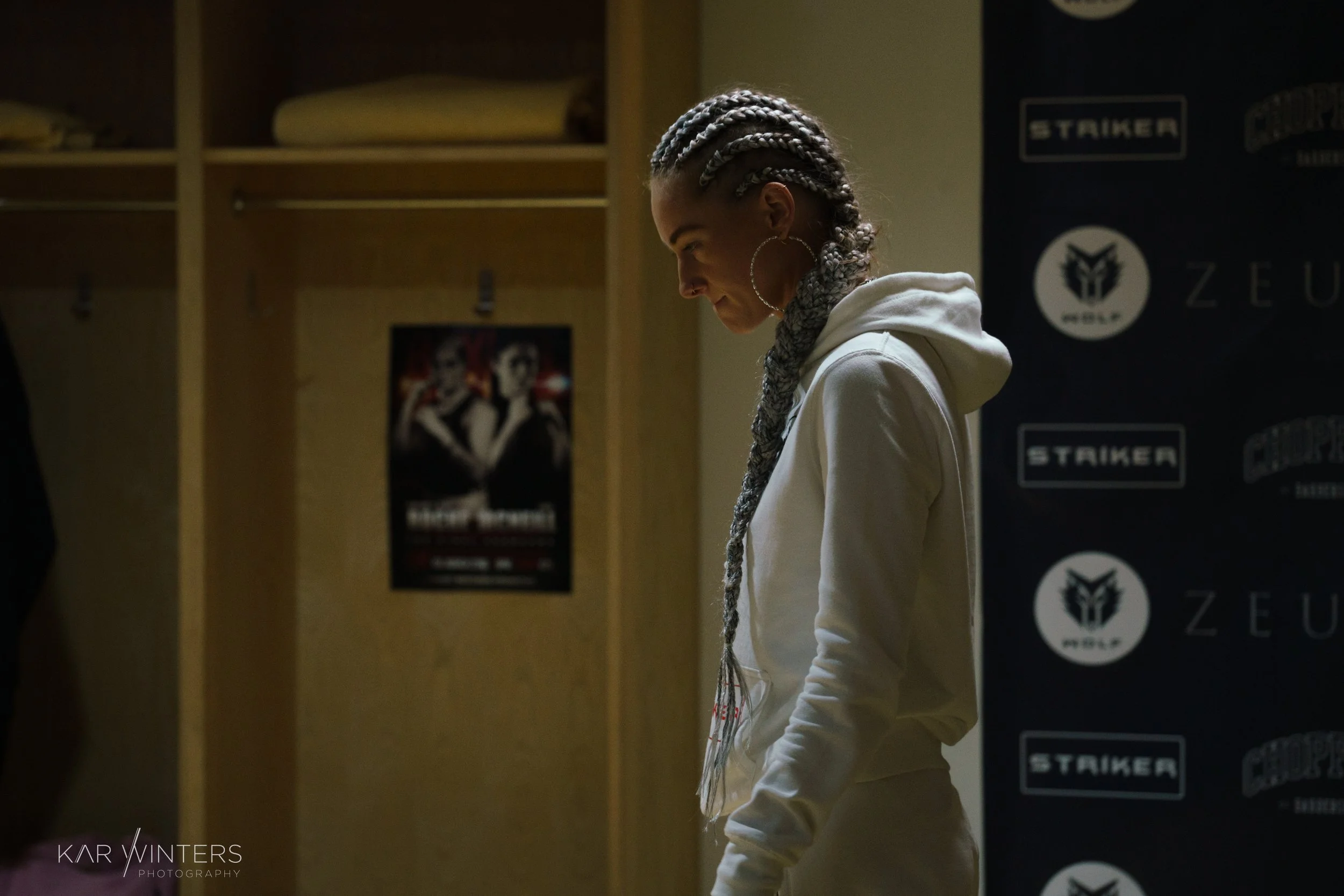 A woman with braided hair, gold hoop earrings, and a gray scarf, wearing a white hoodie, standing with her head tilted down in a dimly lit room.