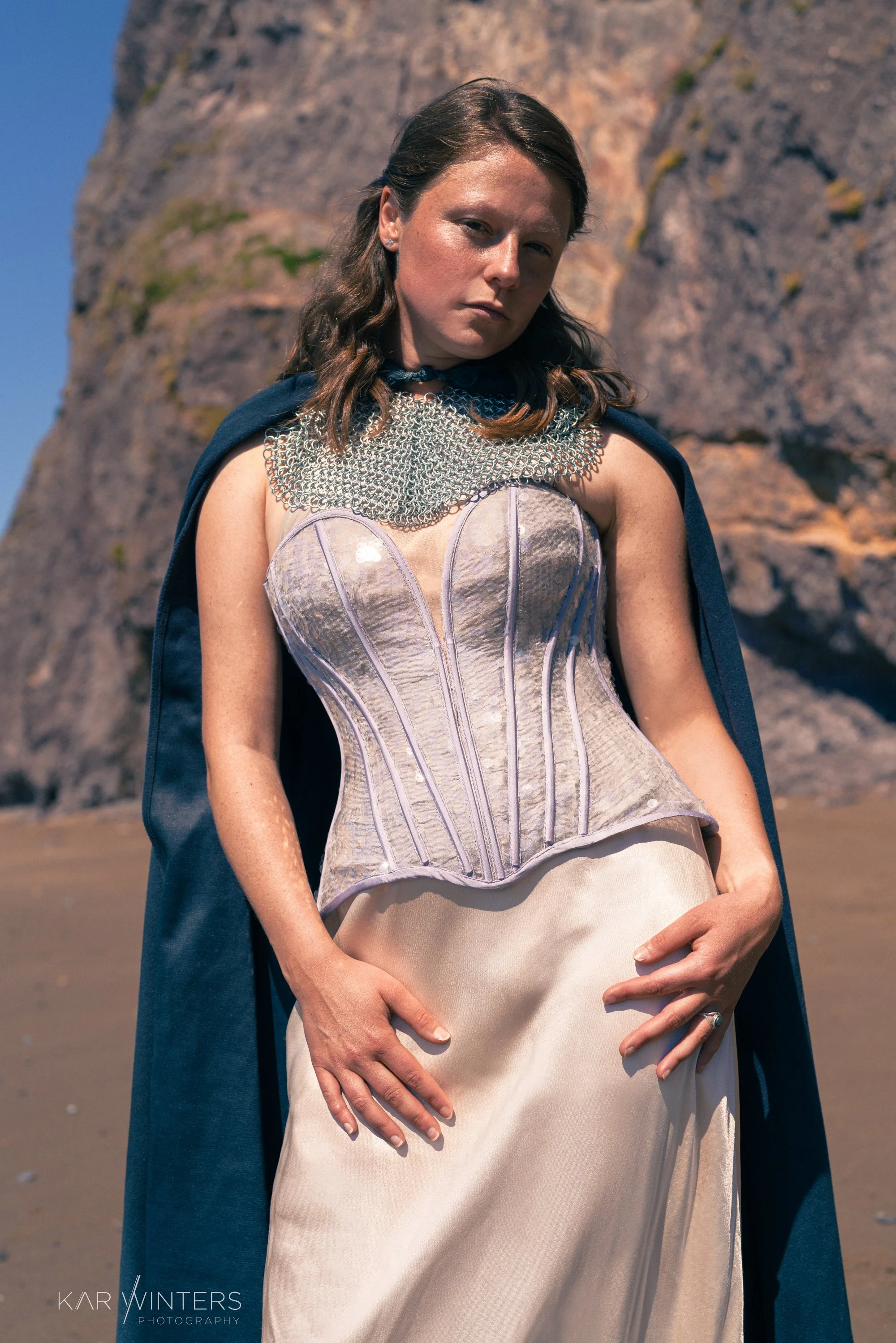 A woman in a medieval-inspired costume with a metallic corset, chainmail neck piece, blue cape, and cream-colored skirt, standing outdoors near rocky cliffs.